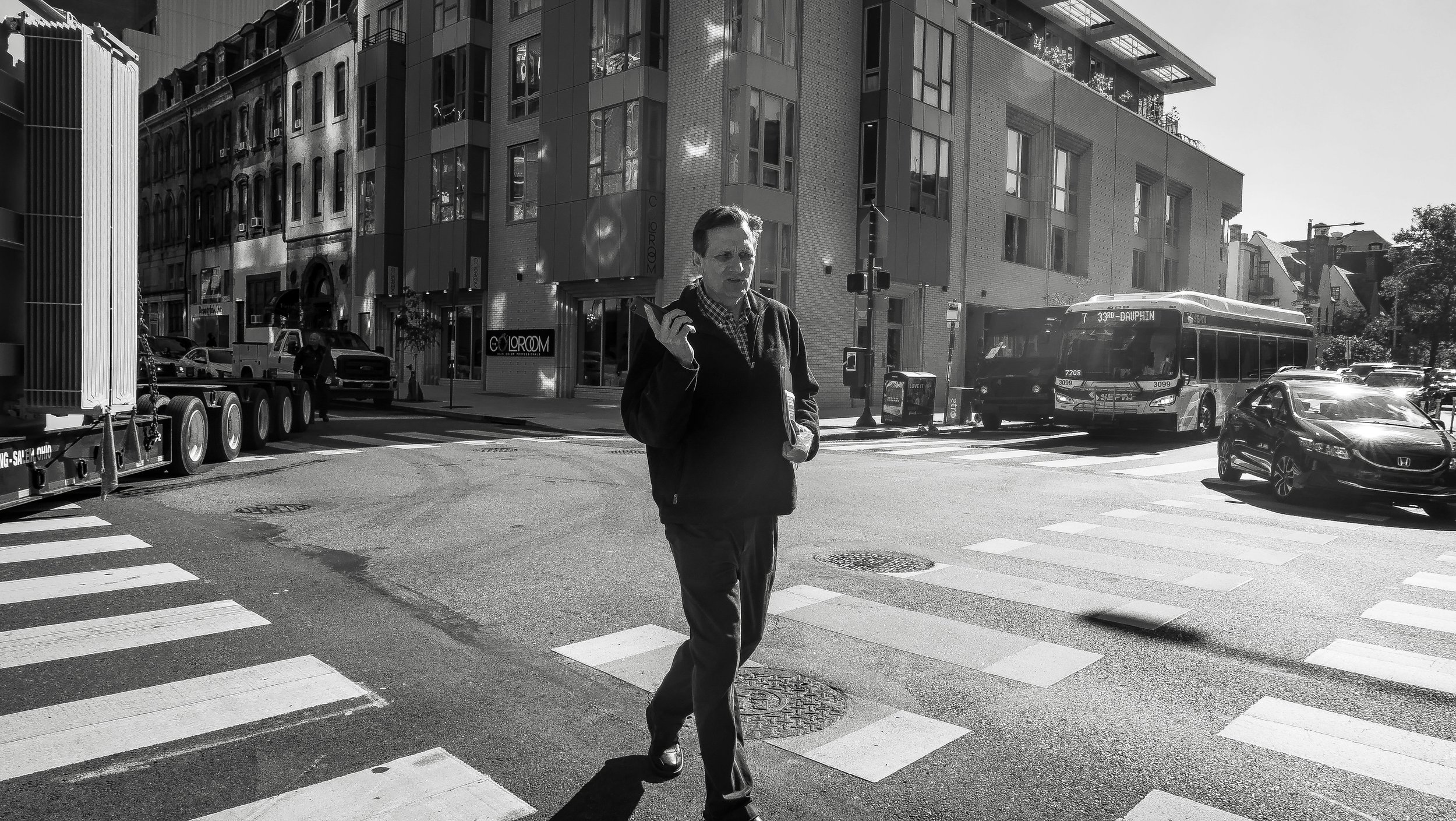 A man walking across a city street with buildings, cars, and a bus in the background, black and white photo.