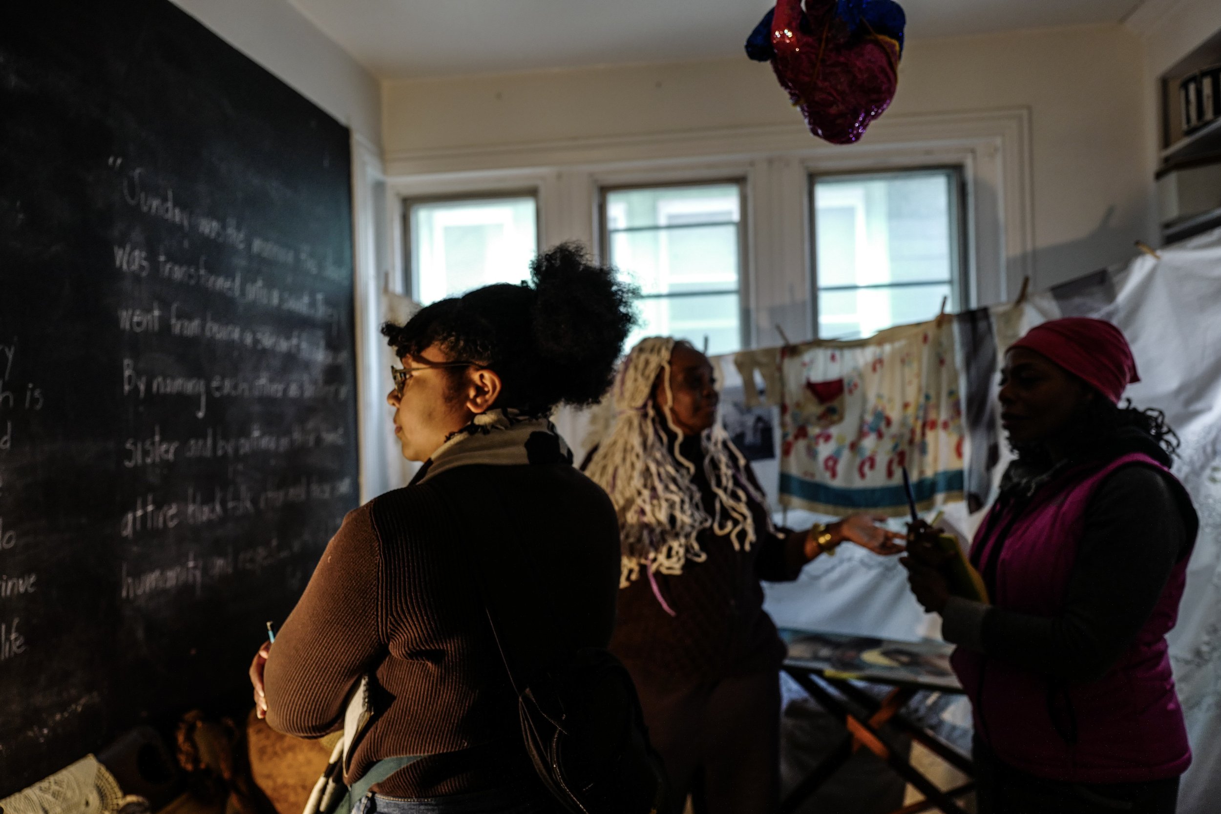 Three women in conversation indoors near a blackboard and window, with laundry hanging in the background.