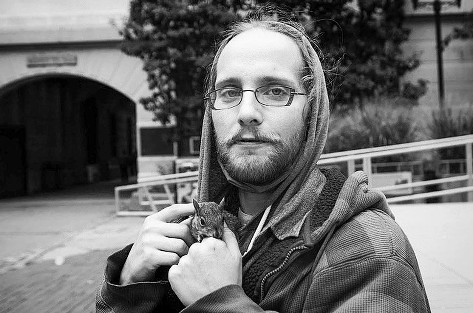 A man with glasses, a beard, and long hair, holding a small squirrel in his hand outdoors in front of a building.