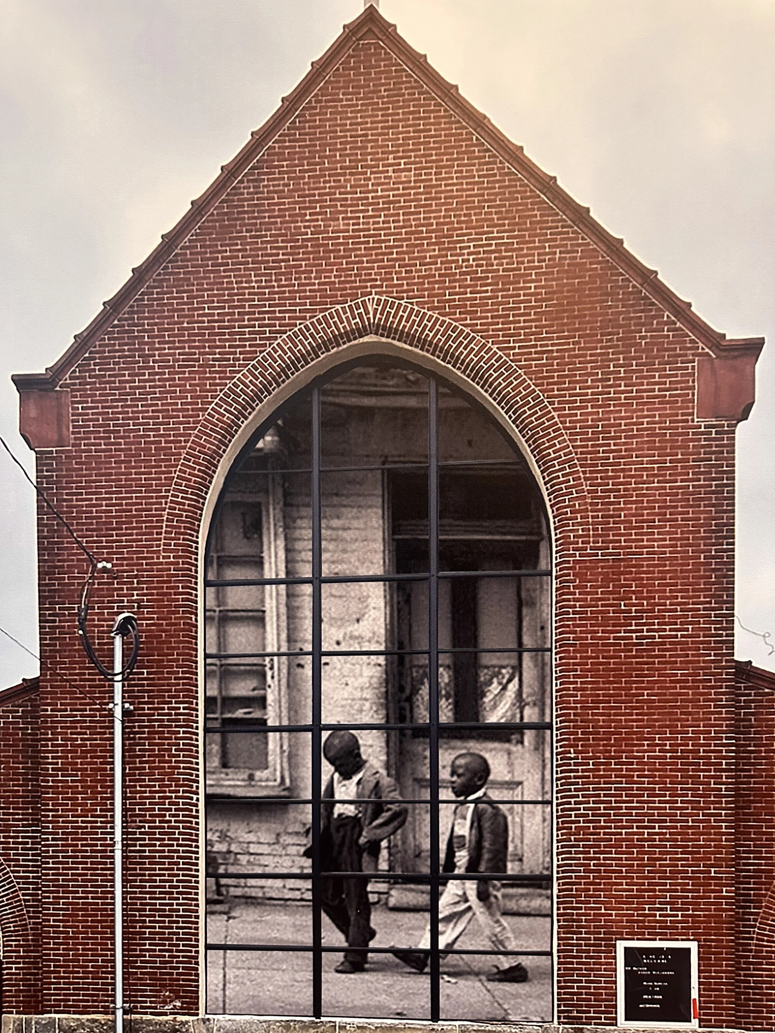 A black and white historical photo of two young boys walking past a decorative brick archway on a city street, with buildings in the background.