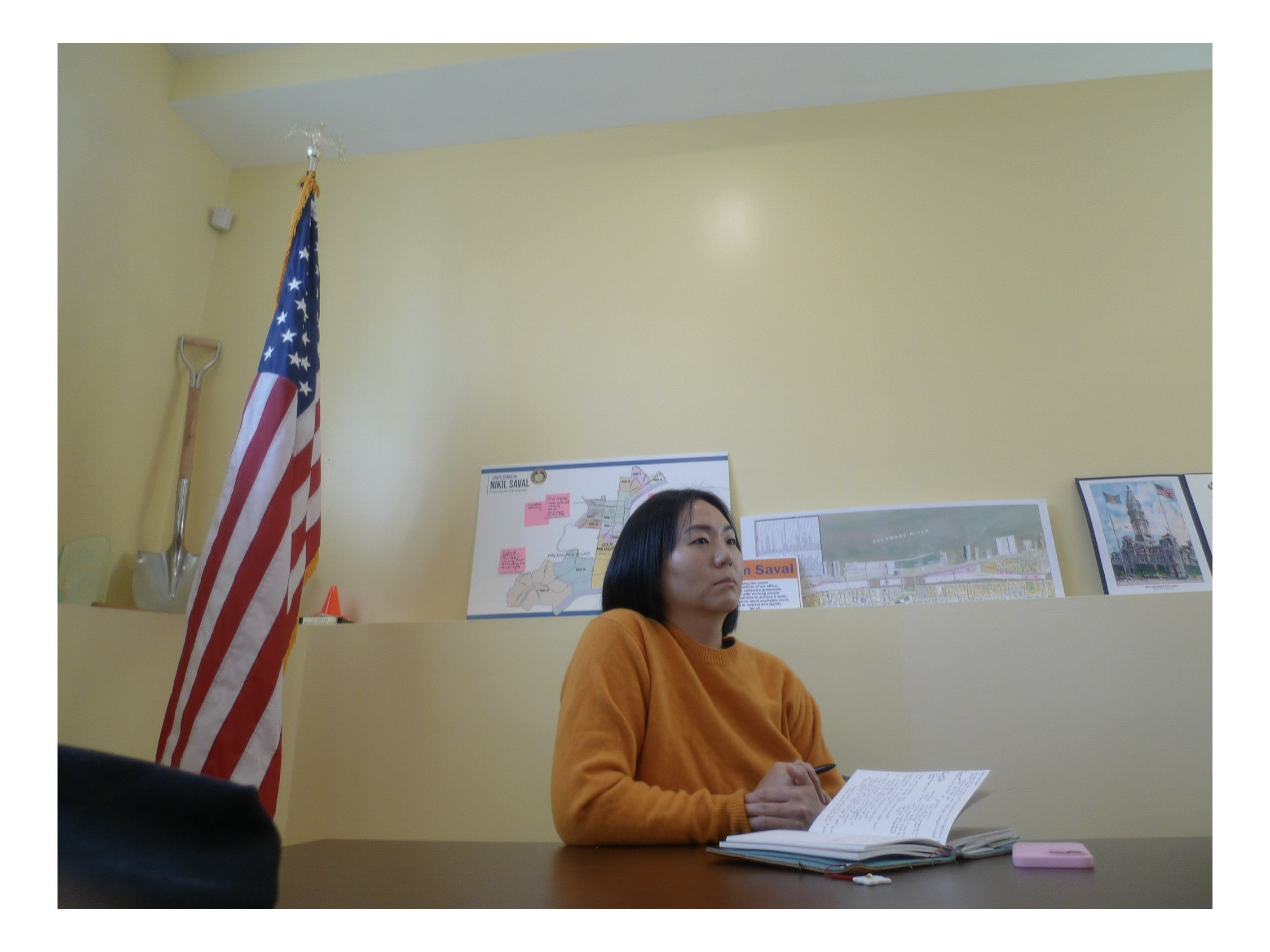 A woman with black hair wearing an orange sweater sitting at a desk, holding papers. Behind her is a yellow wall, a U.S. flag, and a map or poster on the wall.