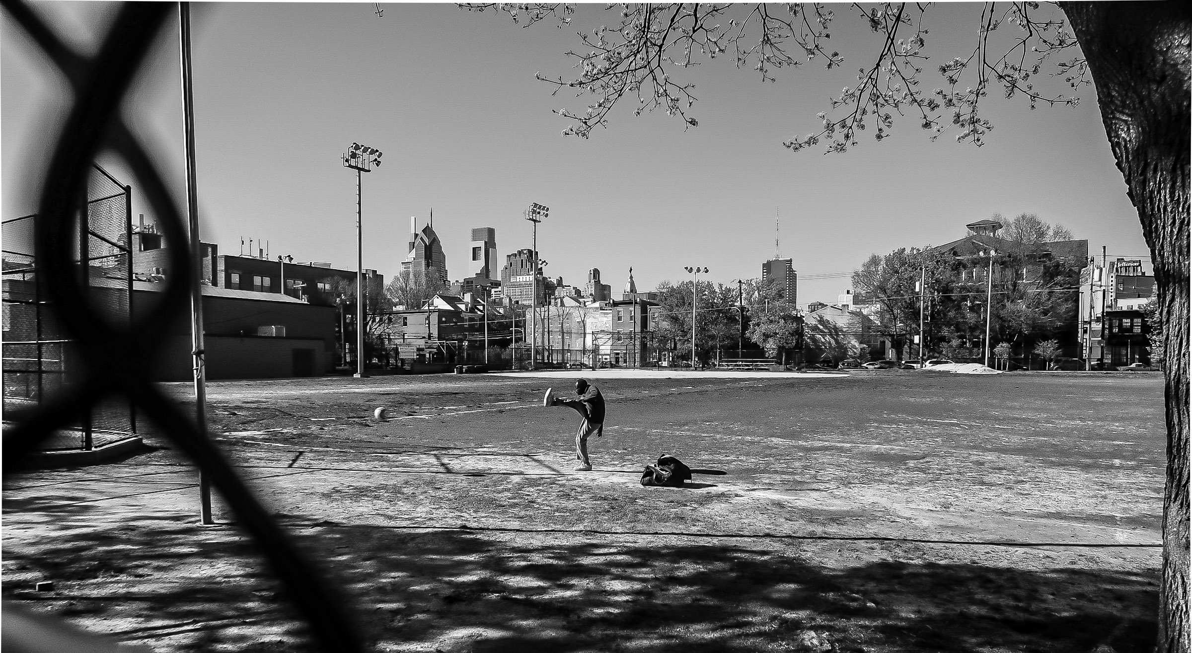 A person is practicing baseball on a baseball field, with a city skyline in the background, viewed through a chain-link fence on a sunny day.