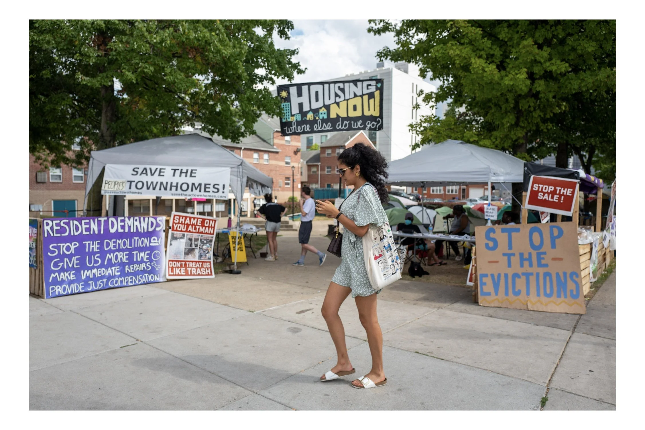A woman with curly hair, sunglasses, and a floral dress walking past protest signs about housing and eviction issues at an outdoor demonstration, with trees and buildings in the background.