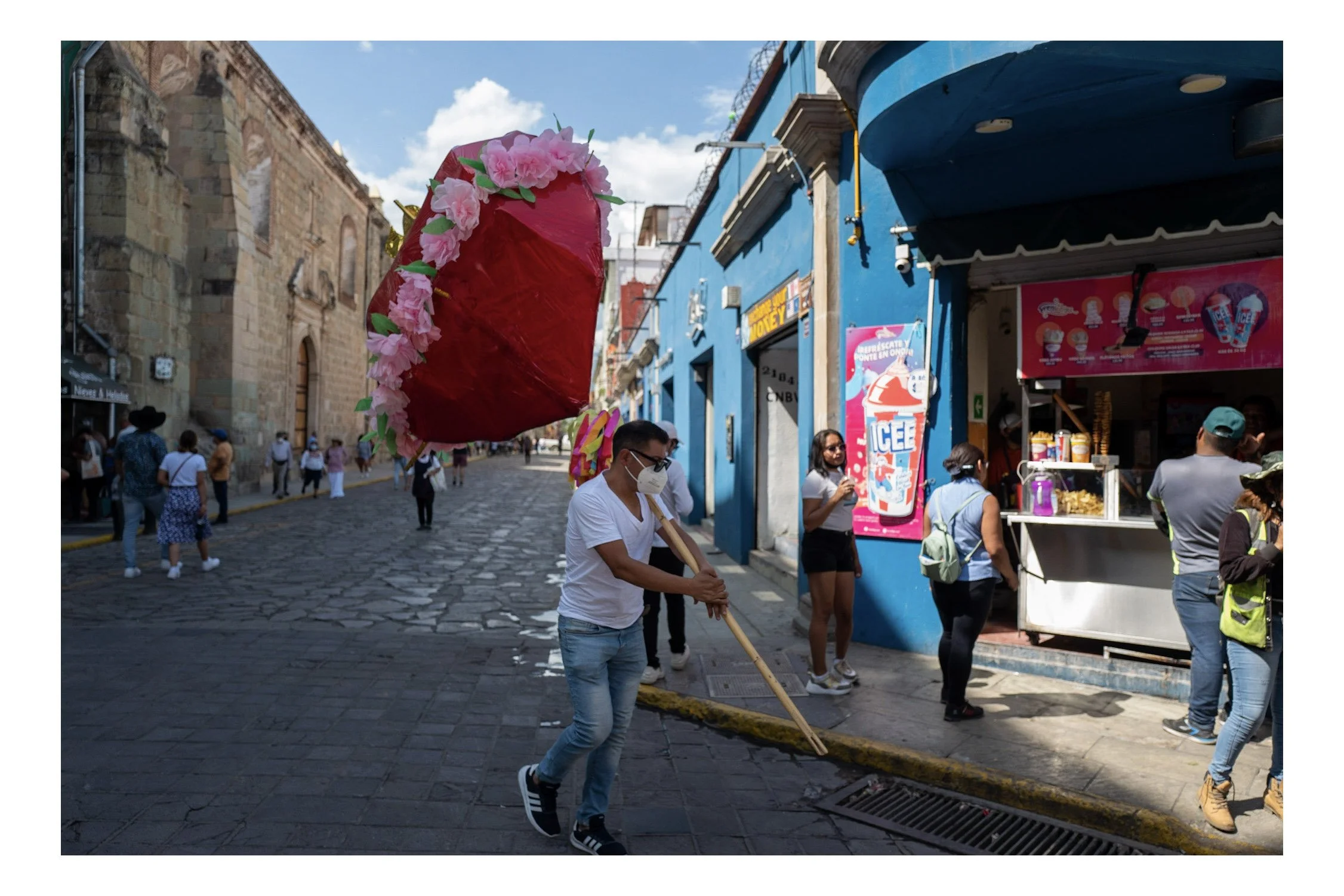 A street scene with a man holding a red piñata decorated with pink flowers while walking among other pedestrians on a cobblestone street. The background features a blue building with signage and a small ice cream stand.