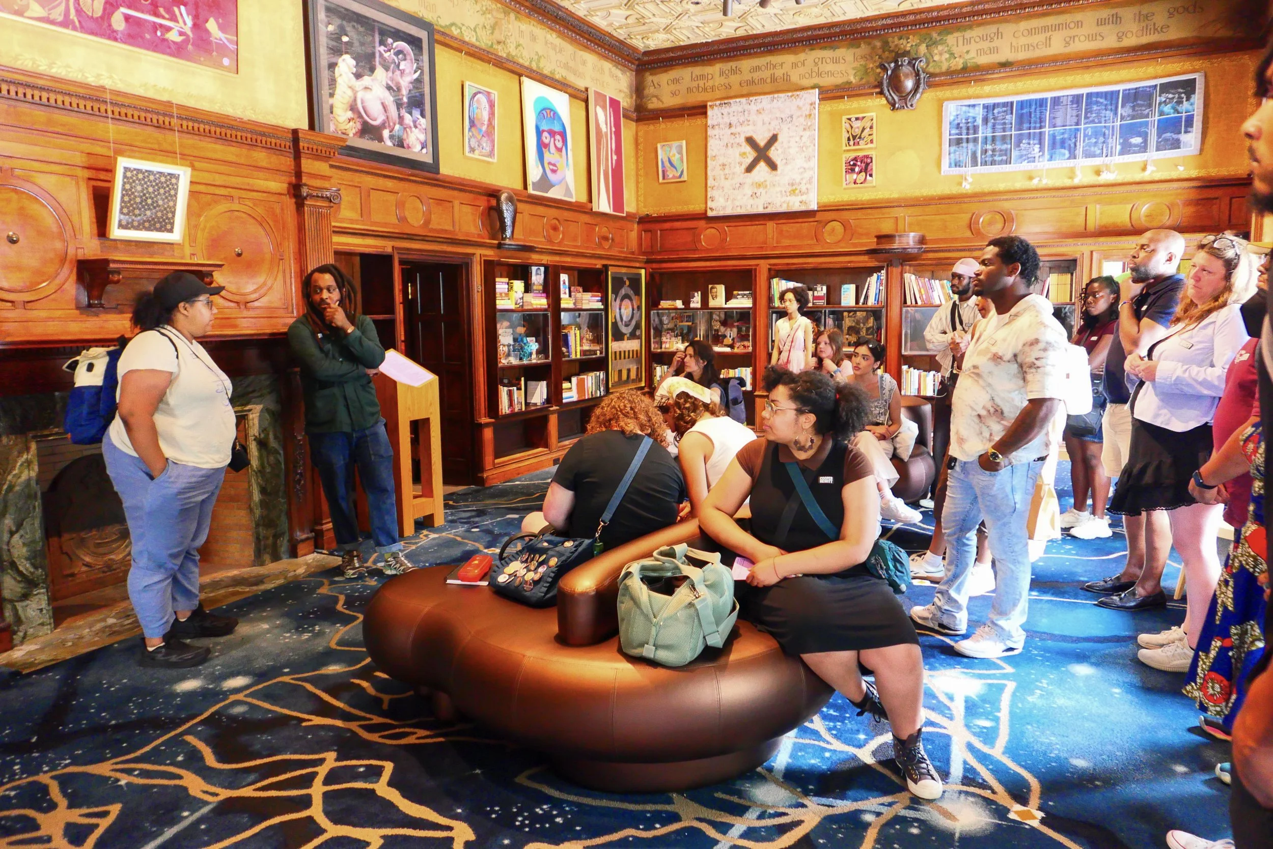 A group of people gathered in a decorated room with wooden shelves filled with books and artwork on the walls. Some people are sitting on a round, brown leather bench, while others are standing. The room has a vibrant blue and gold patterned carpet, 