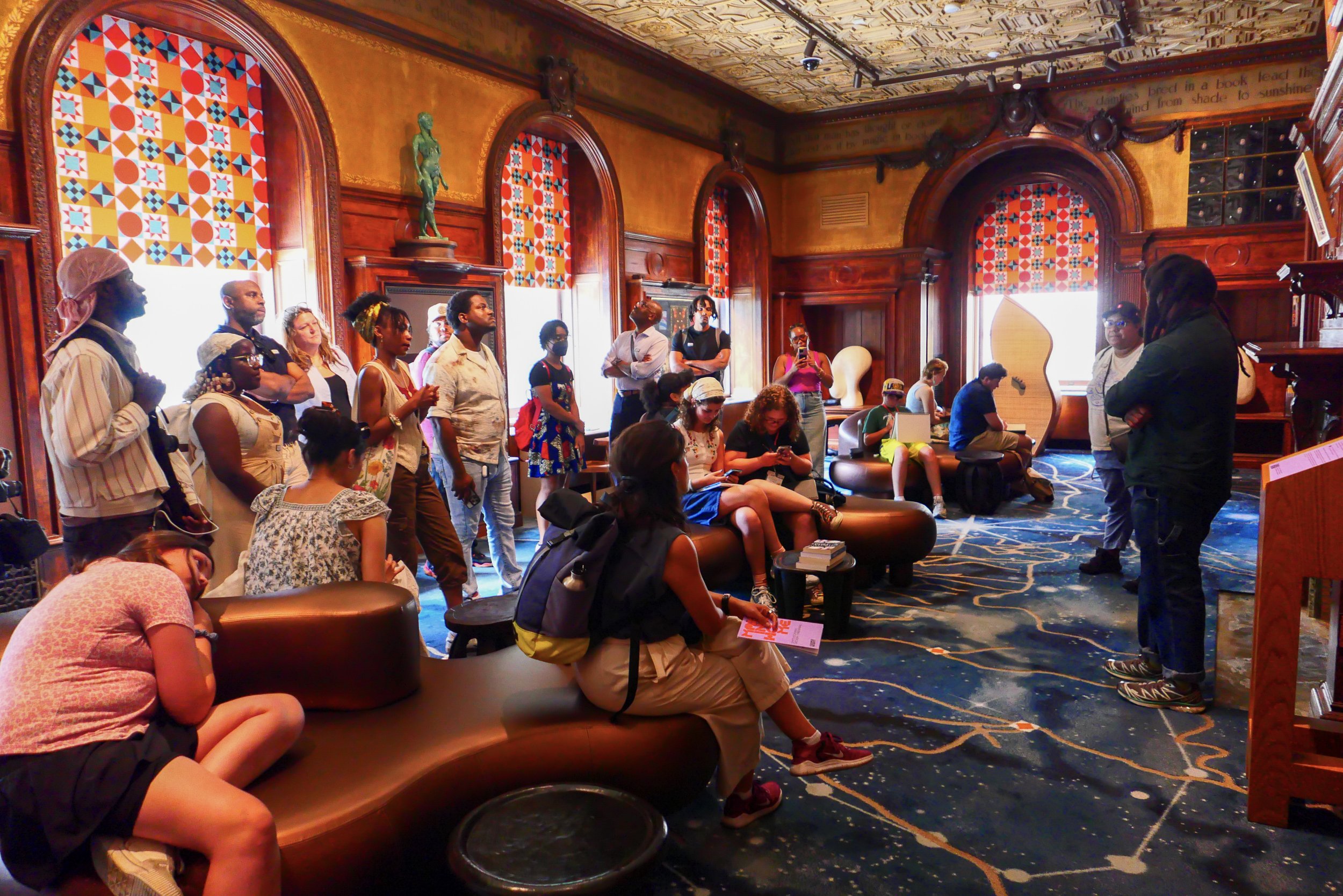 A group of diverse people gathered in a warmly decorated room with rich wood paneling and colorful stained glass windows. Some are seated, others are standing, listening to a speaker at the front. The room features sculptures, a grand piano, and orna