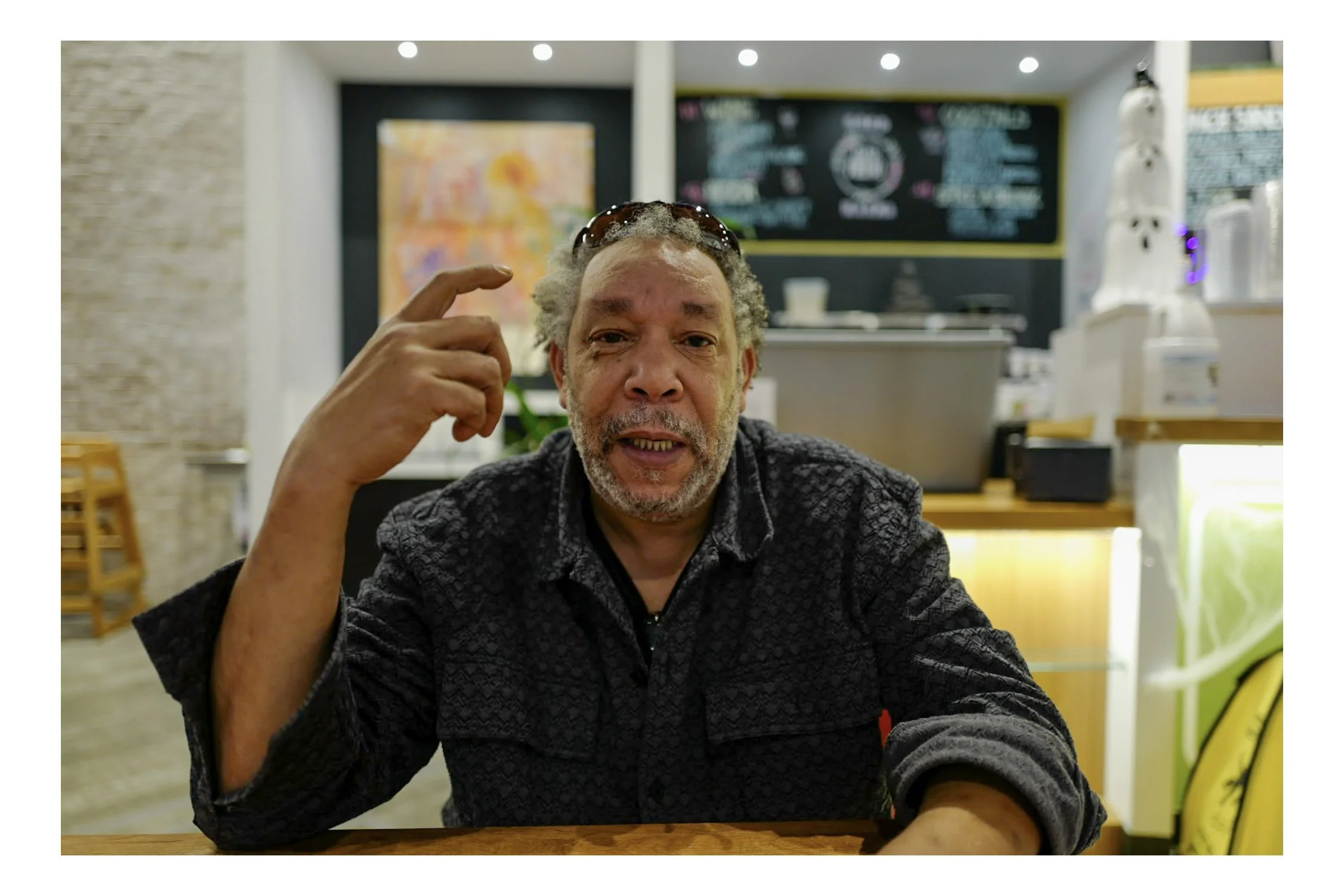 A middle-aged man with gray curly hair and a beard sitting in a cafe, touching his temple with his right hand, wearing sunglasses on his head and a patterned black jacket, with a blurred menu board behind him.