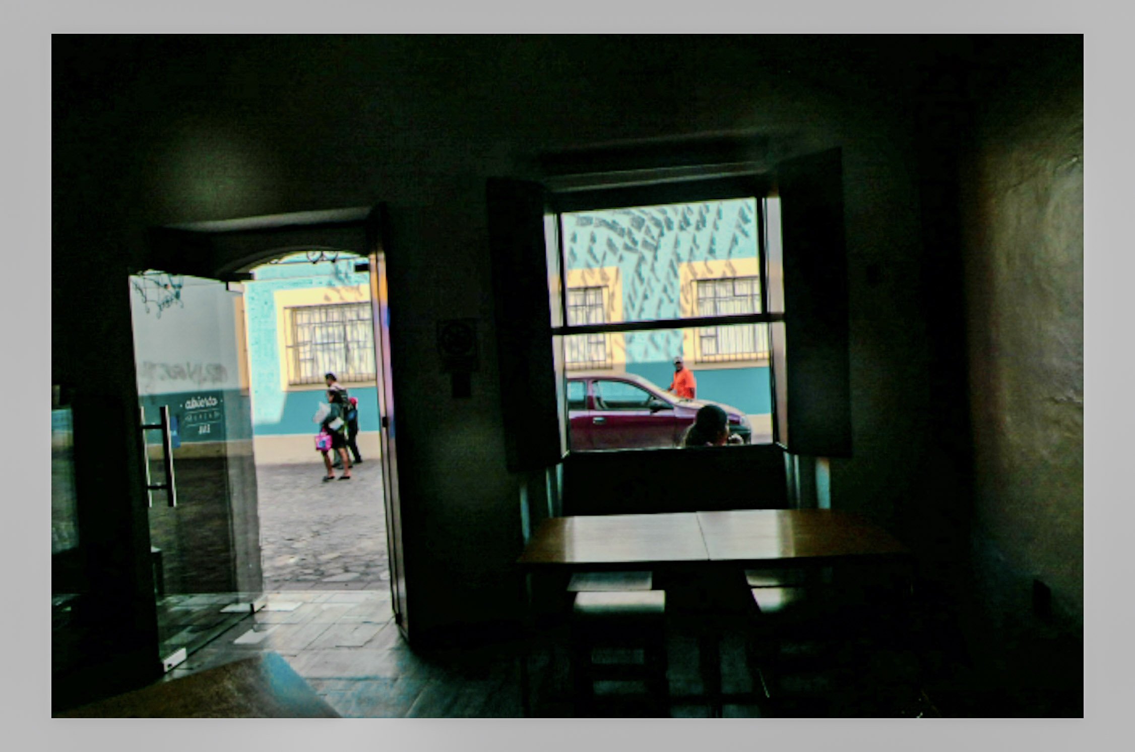 View through a window showing people walking outside, a red car, and colorful buildings with yellow and turquoise walls.