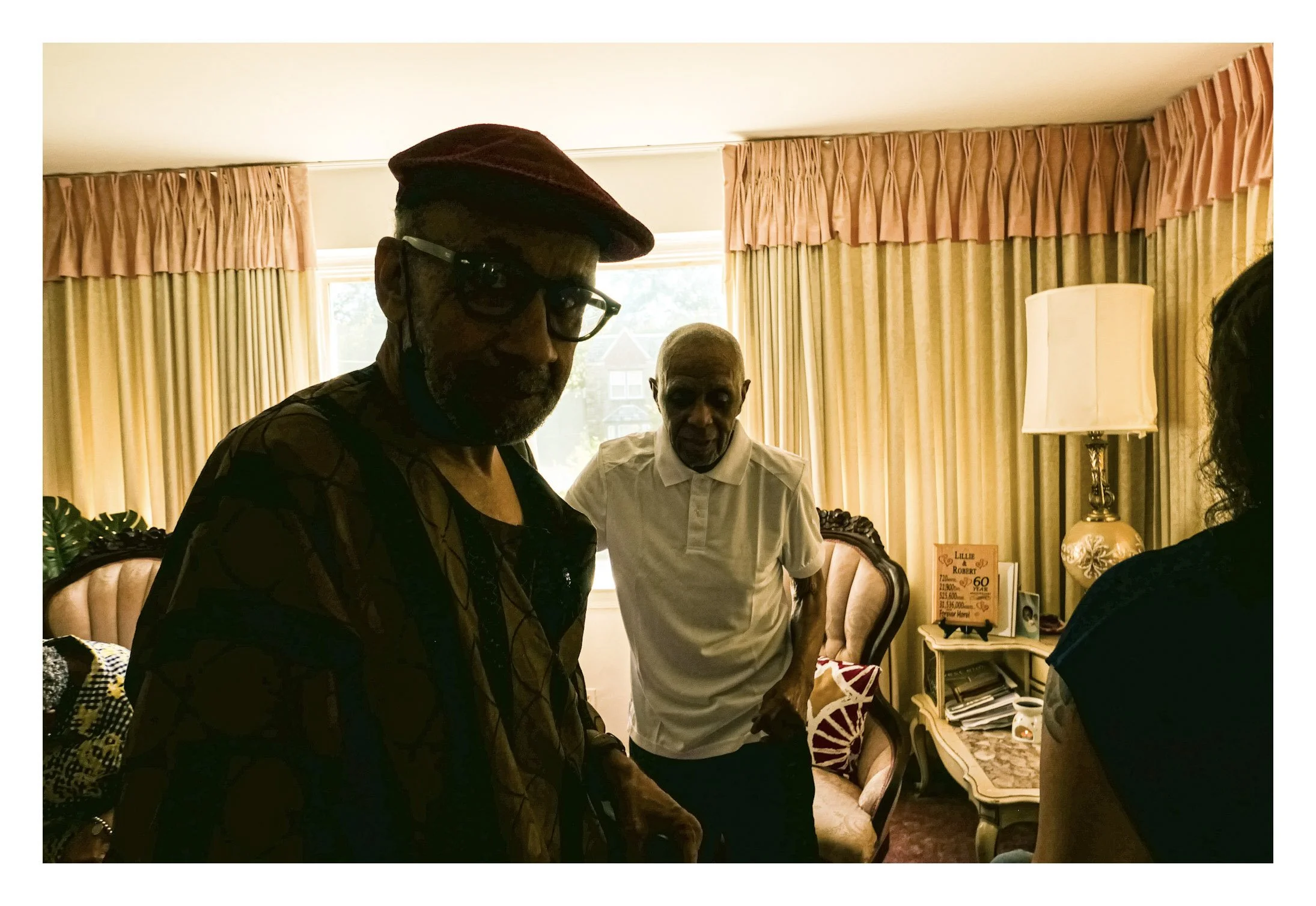 Three men standing in a living room with yellow curtains, a beige lamp, and a pink sign on a table in the background.