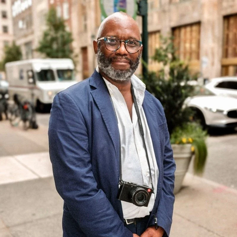 A bearded man wearing glasses, a white shirt, and a blue blazer standing on a city sidewalk with parked cars and buildings in the background. He has a camera hanging around his neck.