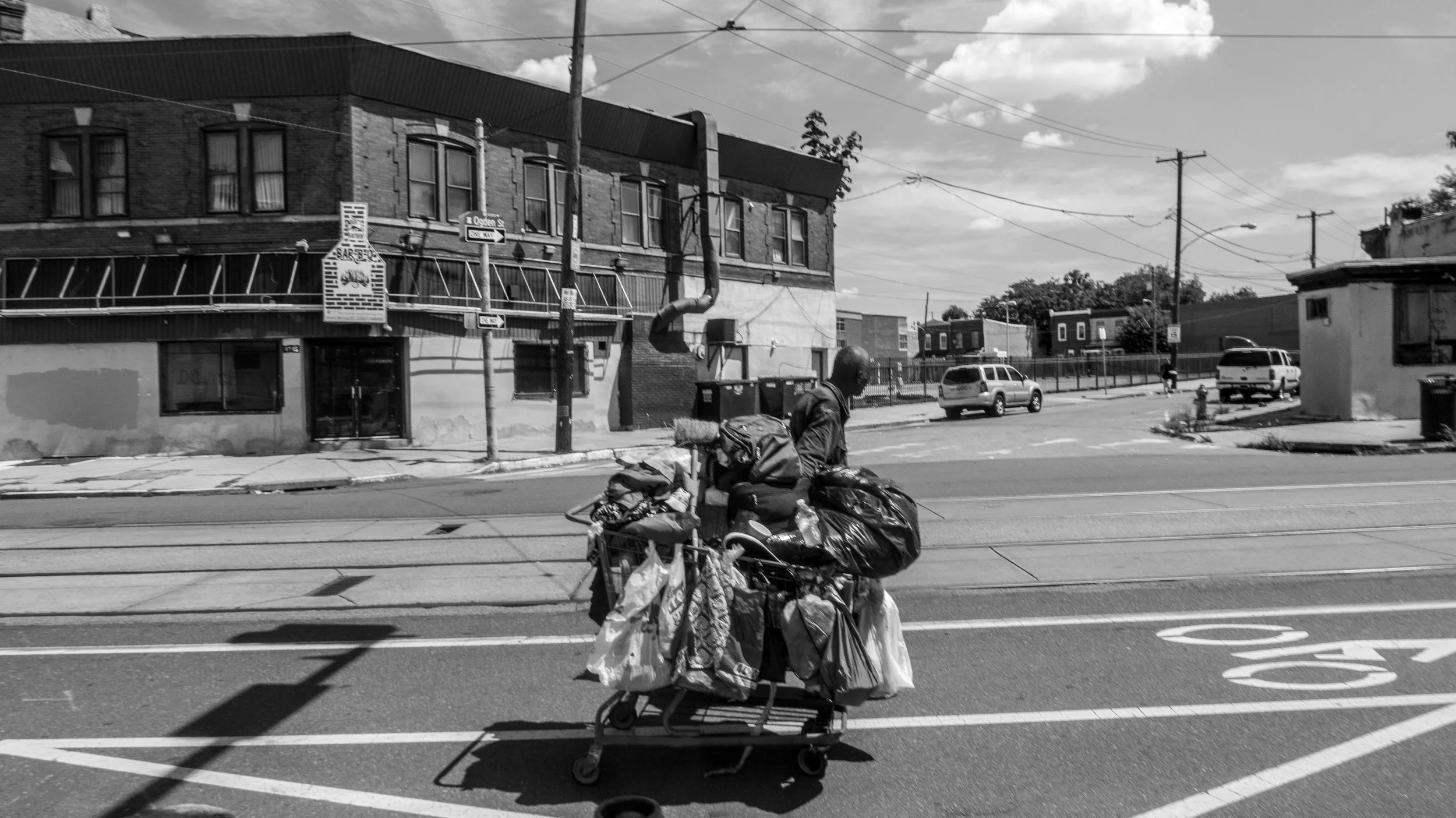 Black and white photo of a man riding a bicycle with several bags attached, crossing a city street with painted tram tracks, in front of a brick building with multiple windows, utility poles, and other buildings in the background, under a partly clou