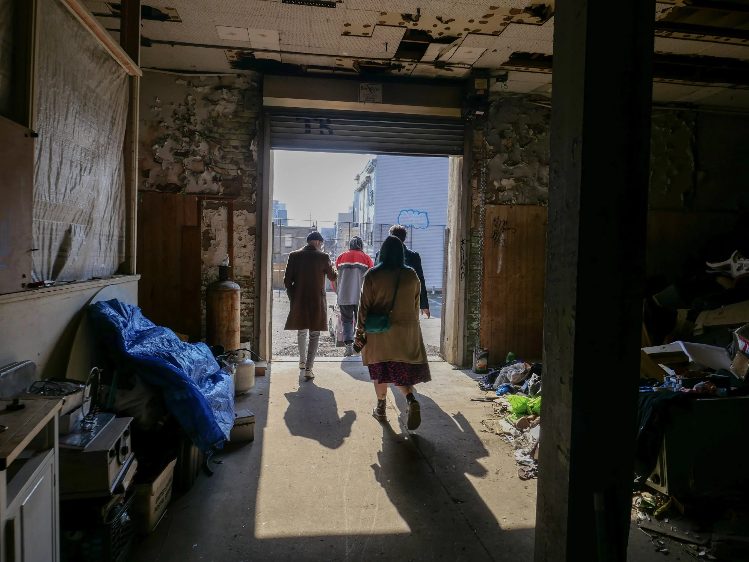 View from inside a building looking out through an open garage door at several people walking outside in an alleyway, with buildings visible in the background.