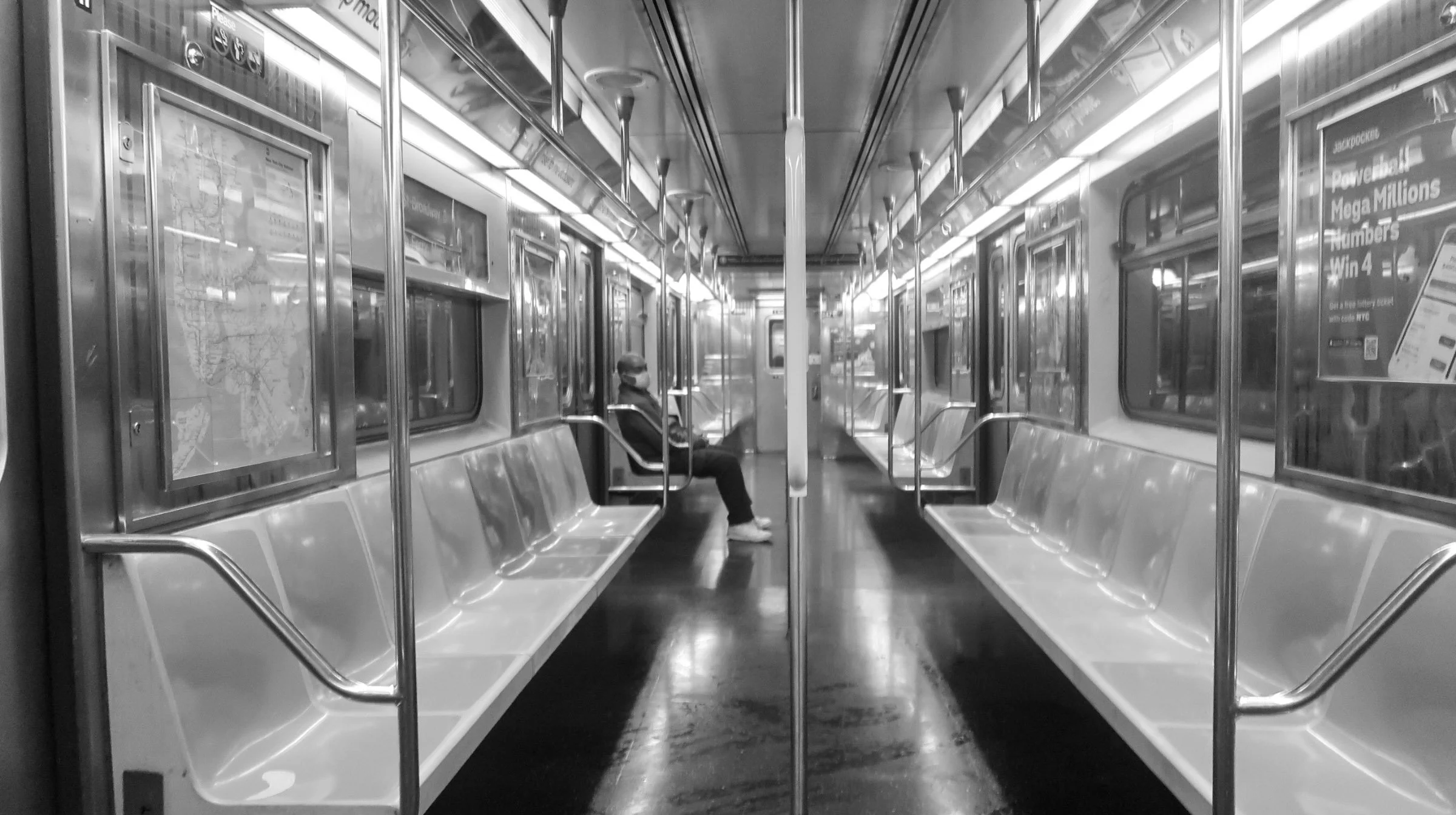 Empty subway car with seats along the sides, a single passenger wearing a face mask sitting on the left side, and subway map and advertisements on the walls.