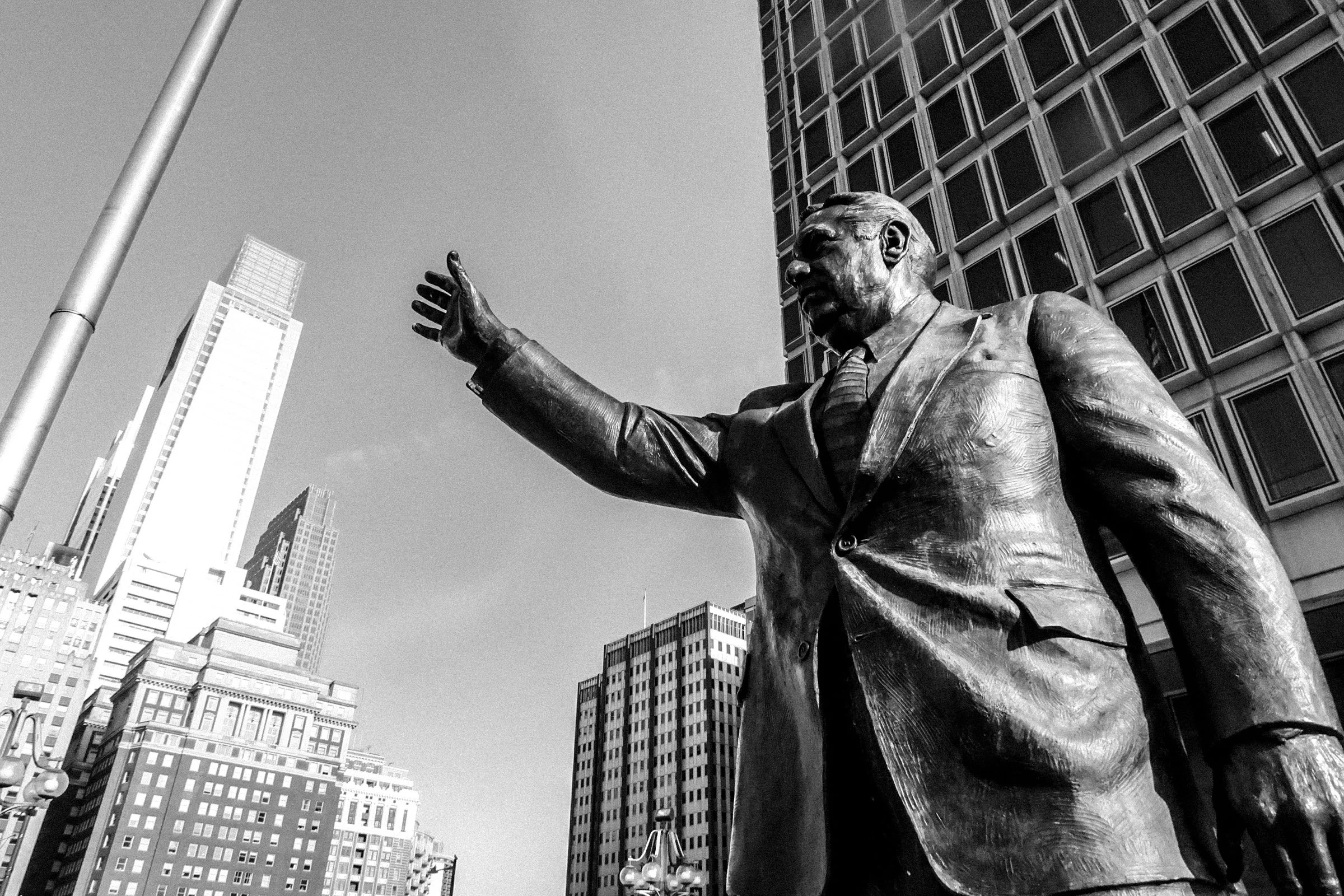 A black-and-white photo of a large statue of a man with a beard, wearing a suit and tie, with his right arm extended outward. The statue is placed in an urban setting with tall buildings and city streets visible behind it.