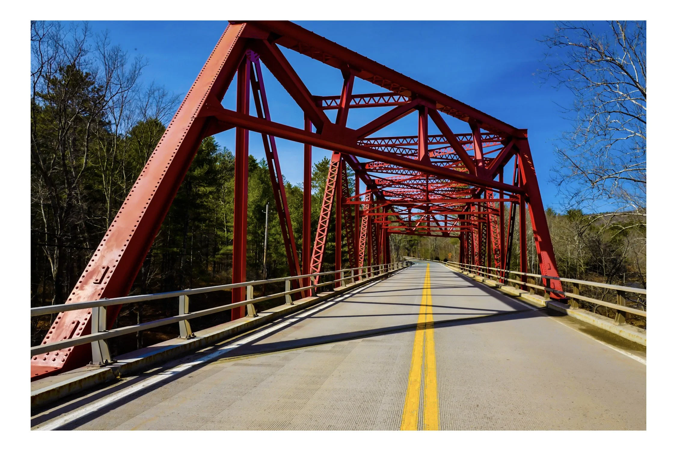 Red truss bridge over a wooded area, with a bright blue sky and no visible vehicles or pedestrians.