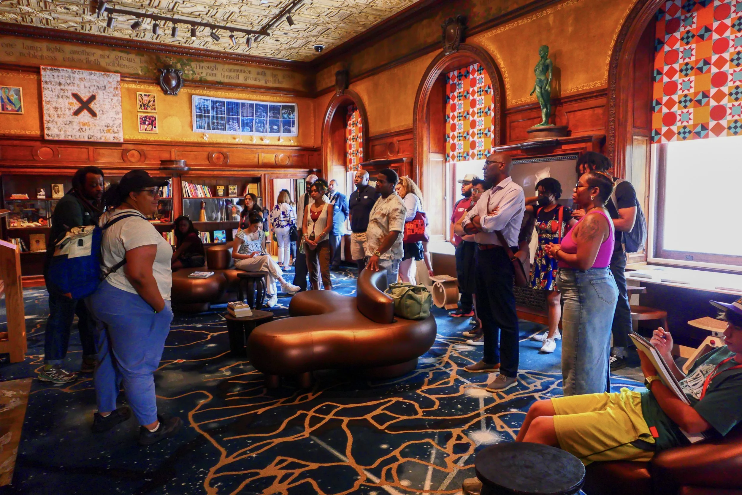 People gathered in a cozy, ornate room with warm orange walls, wooden accents, and colorful geometric patterned window curtains. Some individuals are standing while others are seated, engaged in conversations or listening. The room features bookshelv