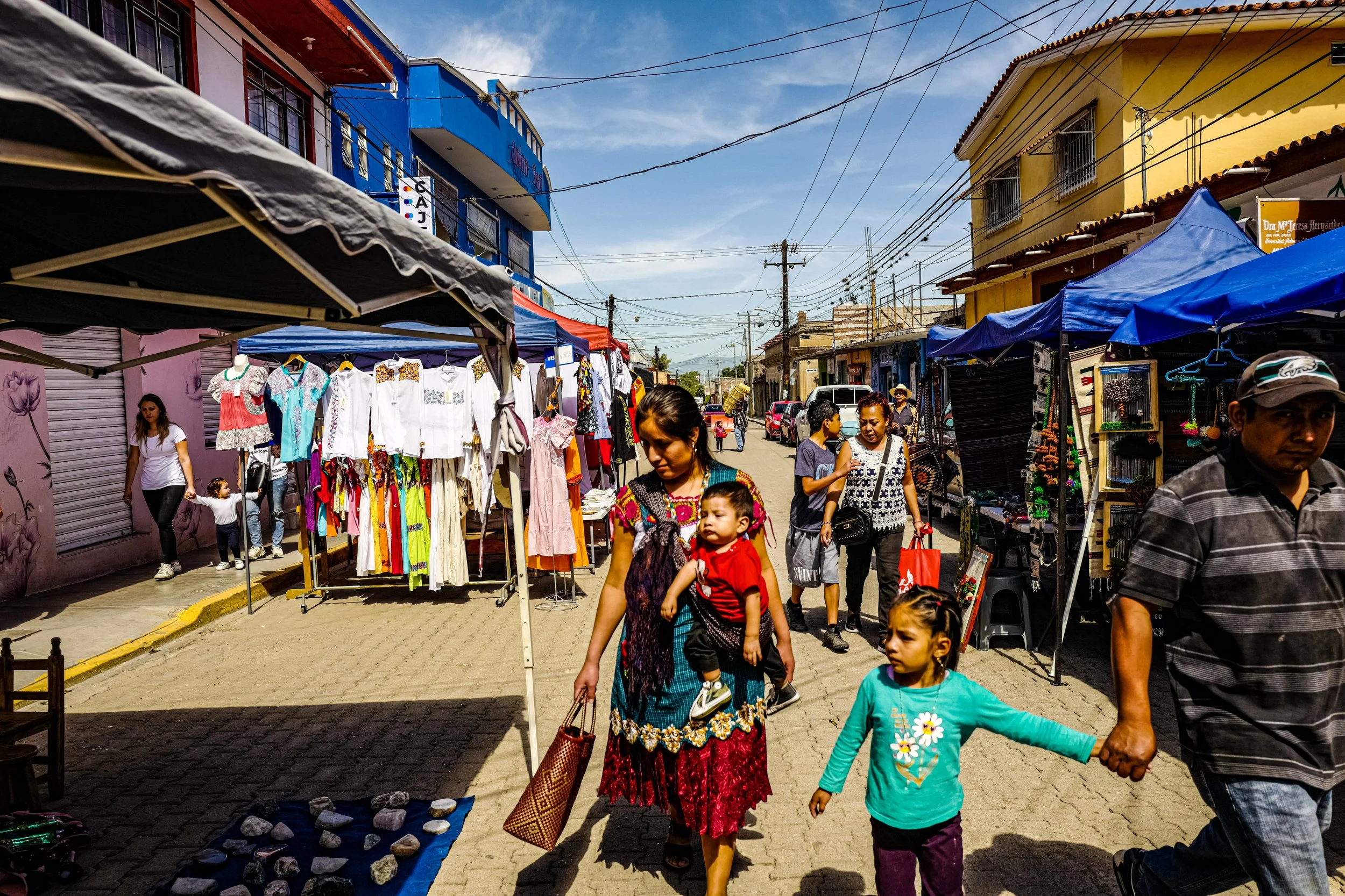 Street market with colorful stalls, selling clothing and jewelry, on a sunny day. People, including children and adults, walk along the street, some holding hands. Buildings are painted in vibrant colors, and electrical wires crisscross in the sky.