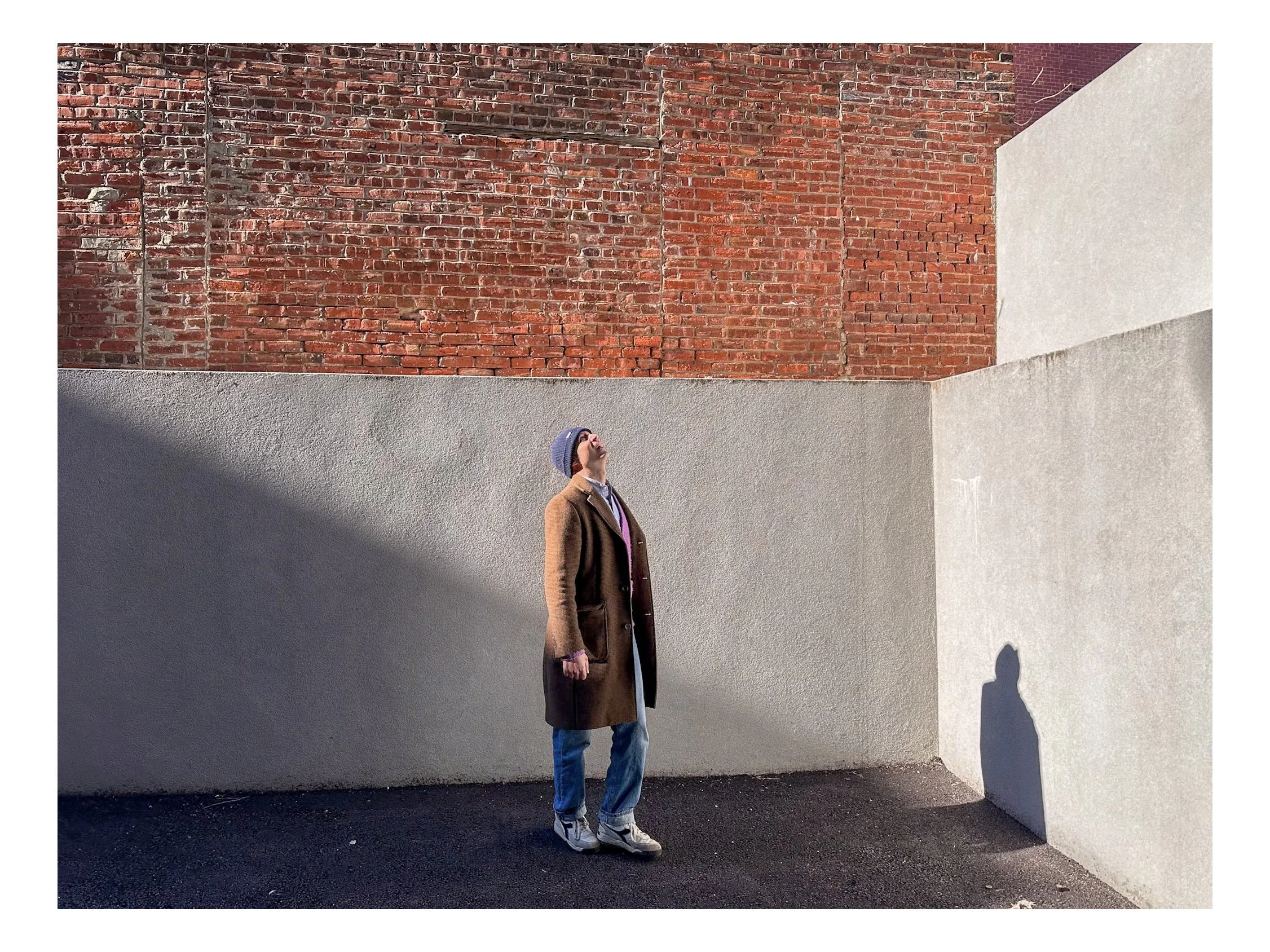A person wearing a brown coat, gray beanie, sunglasses, and sneakers standing against a wall with mixed brick and concrete surfaces, with their shadow cast on the wall.