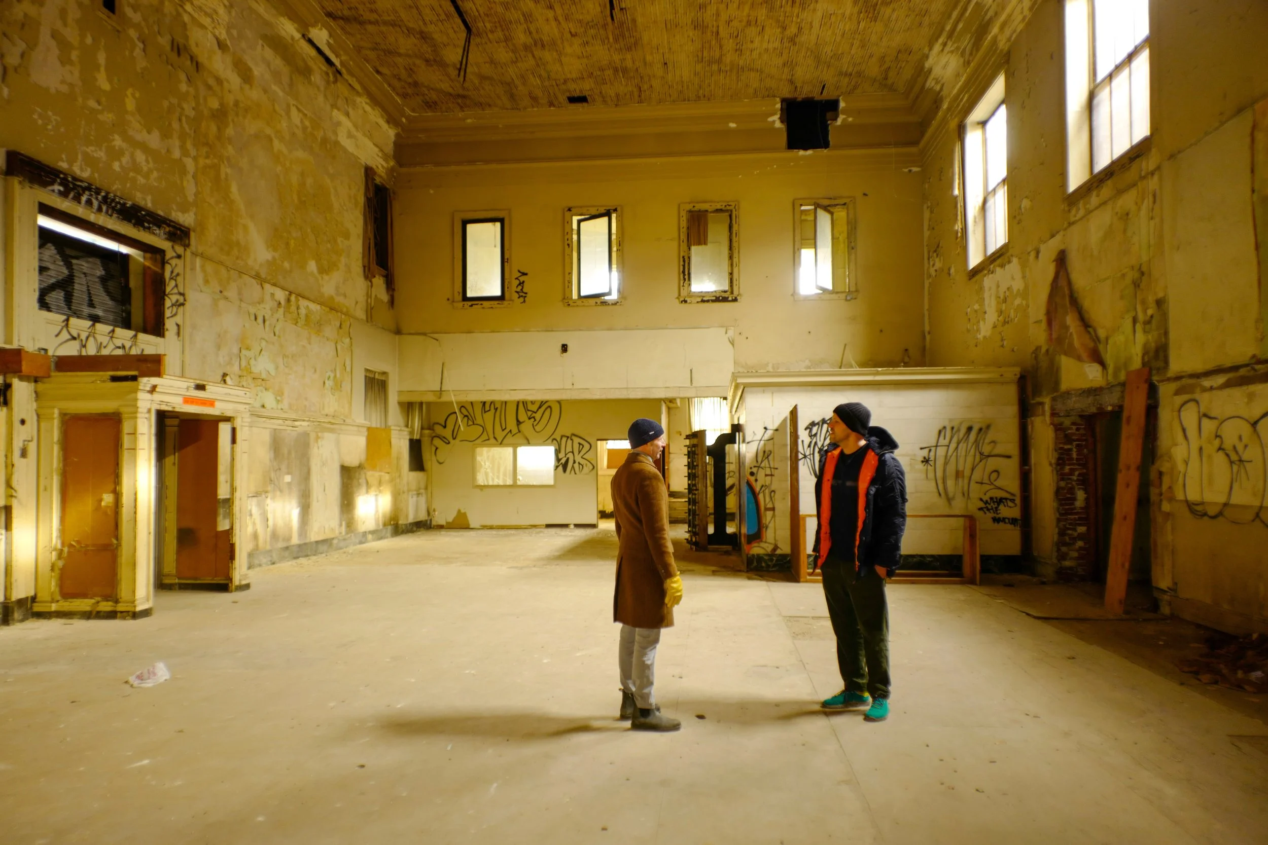 Two people talk inside an old, dilapidated room with graffiti on the walls and broken windows.