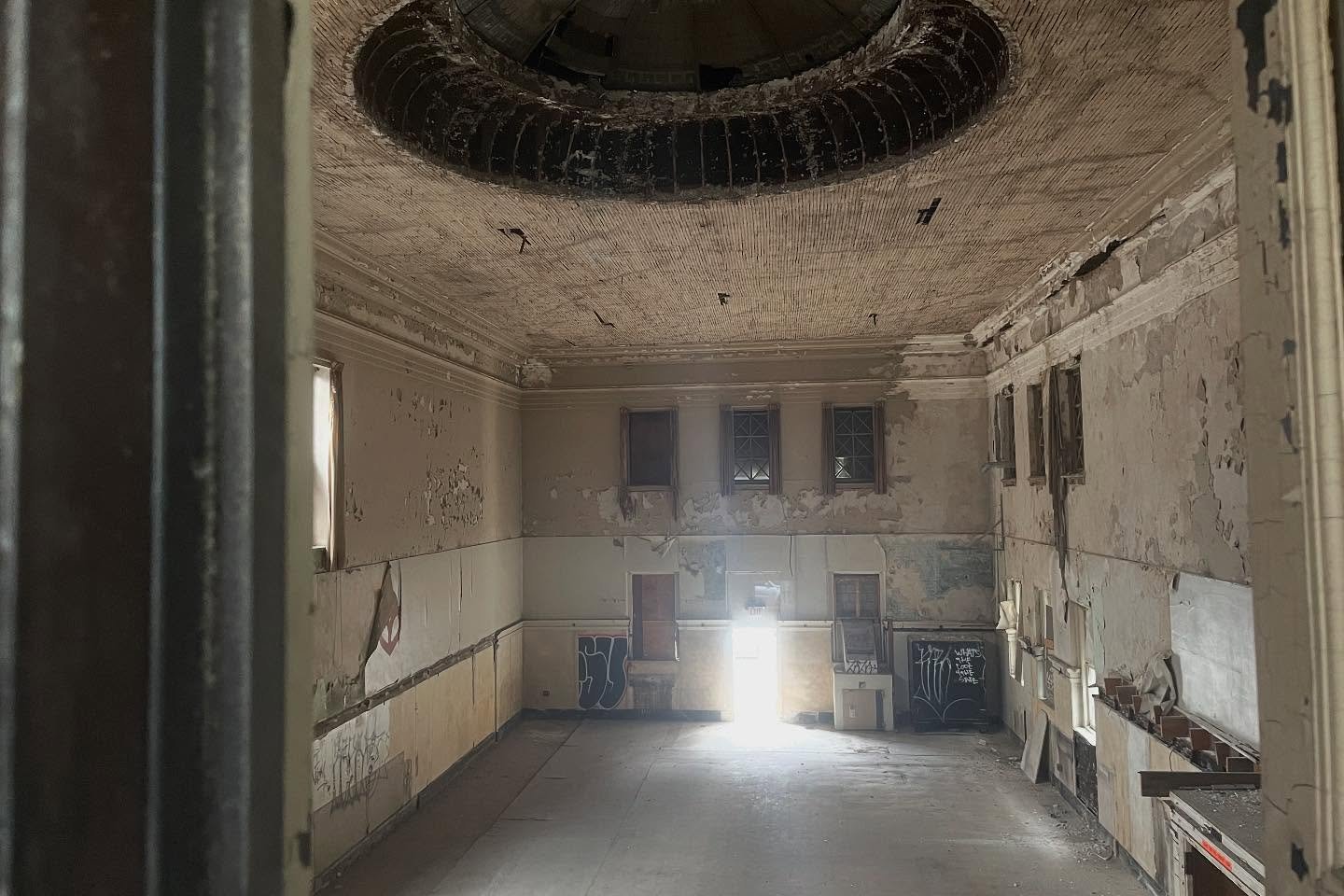 An abandoned, dilapidated room with peeling paint, broken windows, and graffiti on the walls. The ceiling has missing and damaged sections, revealing some structural elements. Light streams in through a door at the far end of the room.