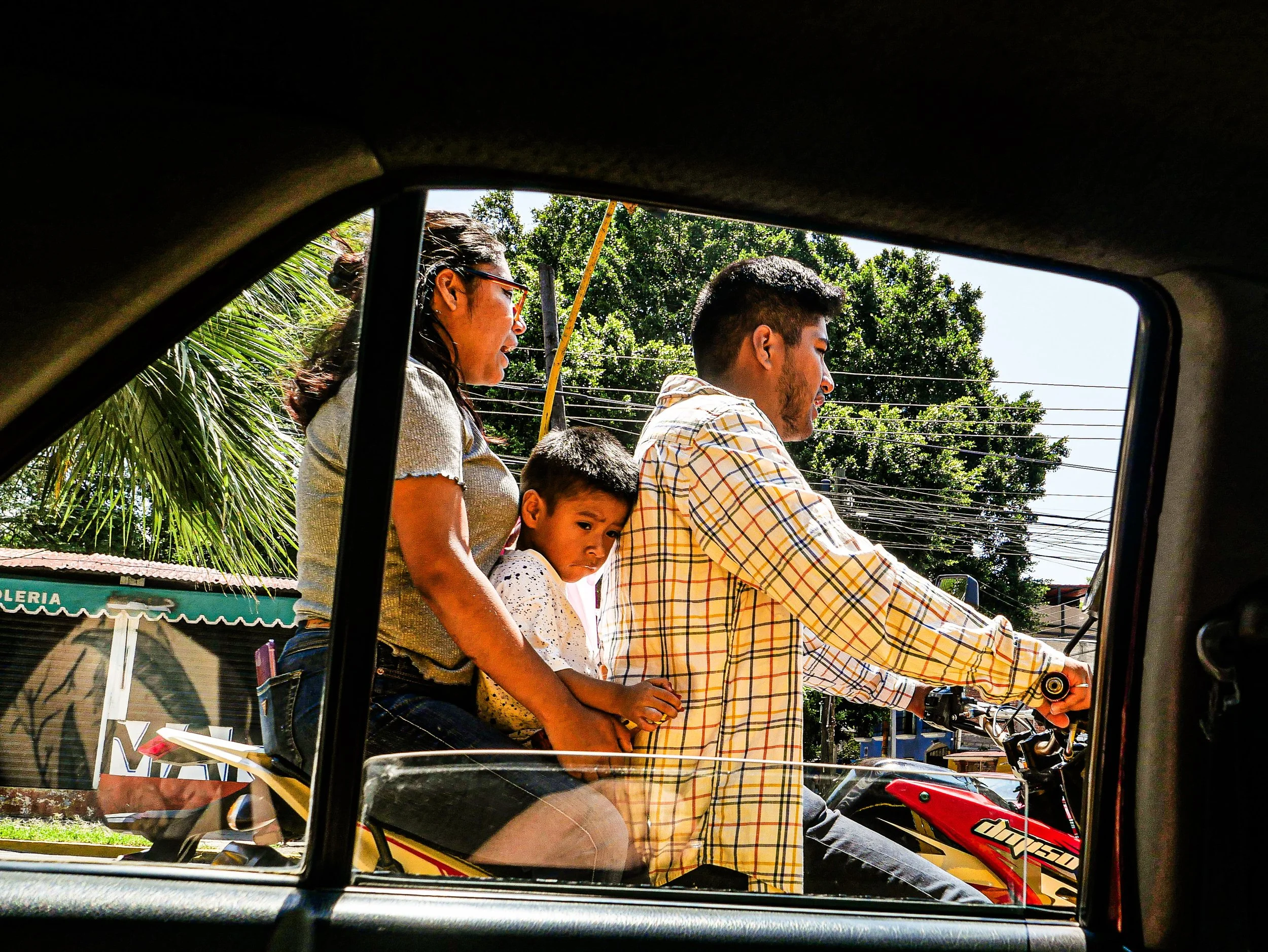 Three people riding a motorcycle, viewed from inside a vehicle, with trees and utility poles in the background.