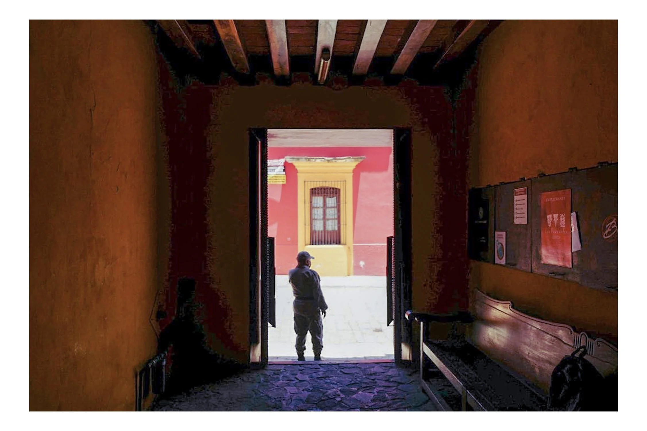 View from inside a dimly lit corridor looking towards an open doorway revealing a square with a person standing outside. The person is wearing a cap and gray clothing. The corridor has orange walls, wooden beams on the ceiling, and benches on the rig
