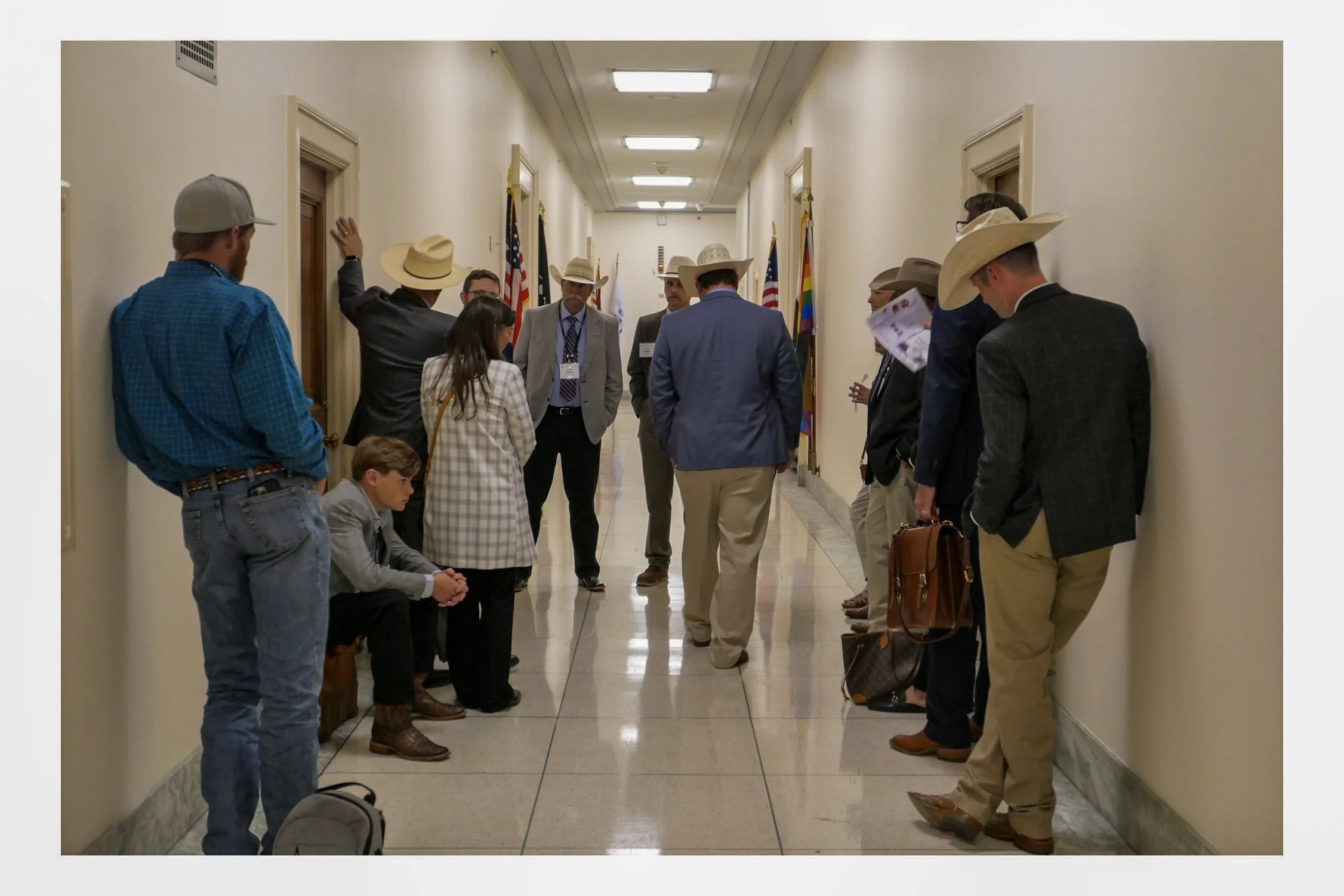 People gathered in a hallway, some standing and others sitting, with American flags and a rainbow flag in the background, participating in a discussion or waiting.