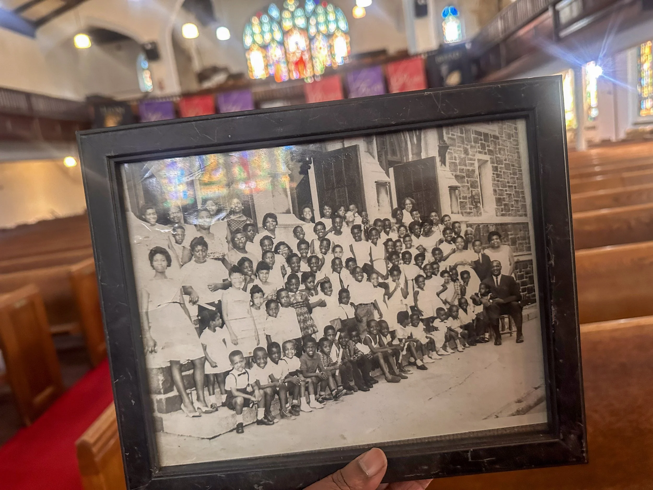 Black and white group photo of children and adults standing and sitting outside a brick building.