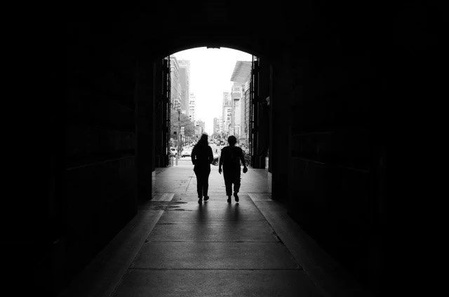 Two people walking through an arched tunnel on a city street in black and white.