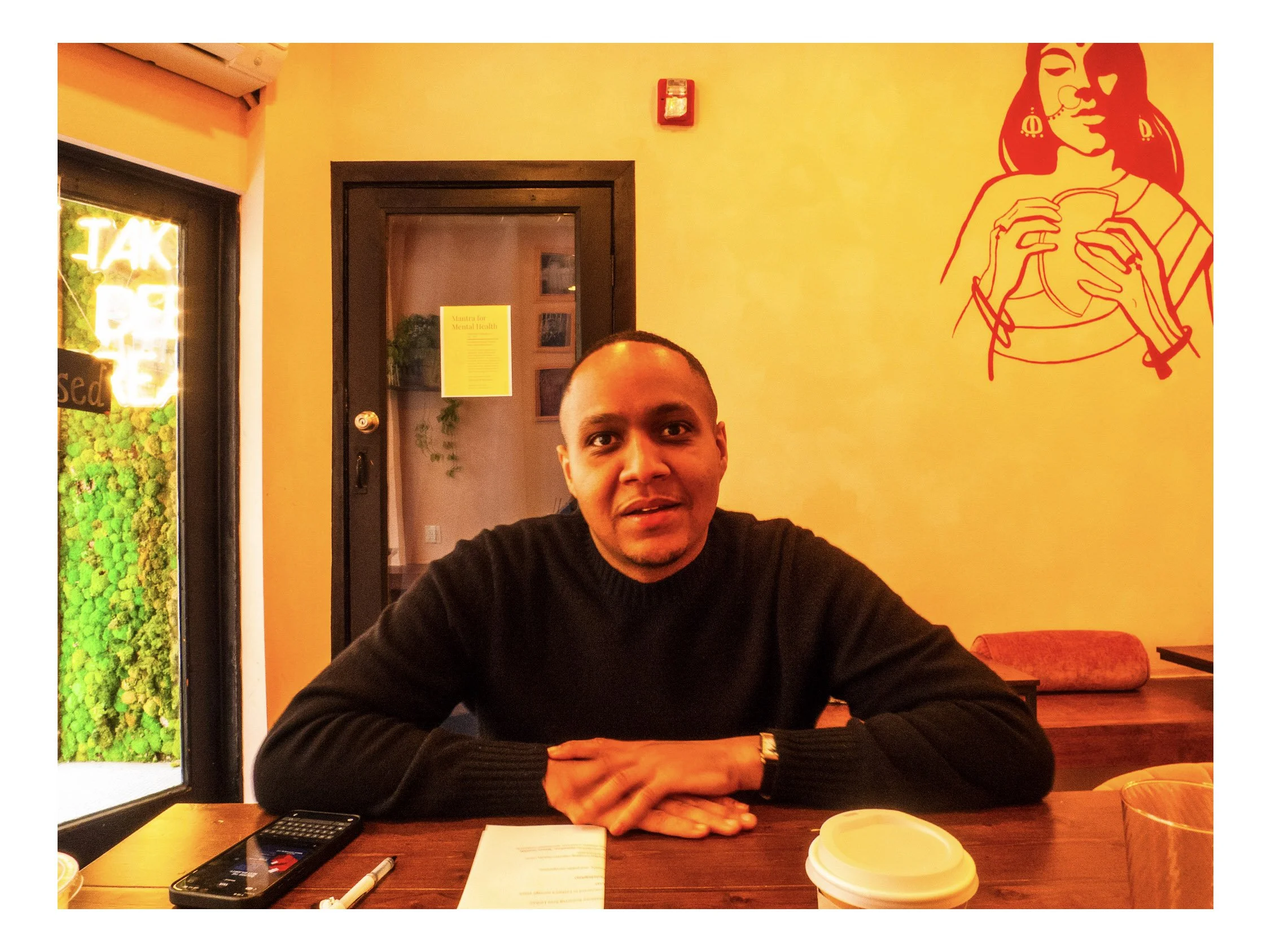 A man sitting at a wooden table in a brightly lit cafe, looking at the camera with a slight smile. On the table are a paper, a calculator, and a drink in a paper cup with a lid. Behind him is a yellow wall with a large red line drawing of a woman hol