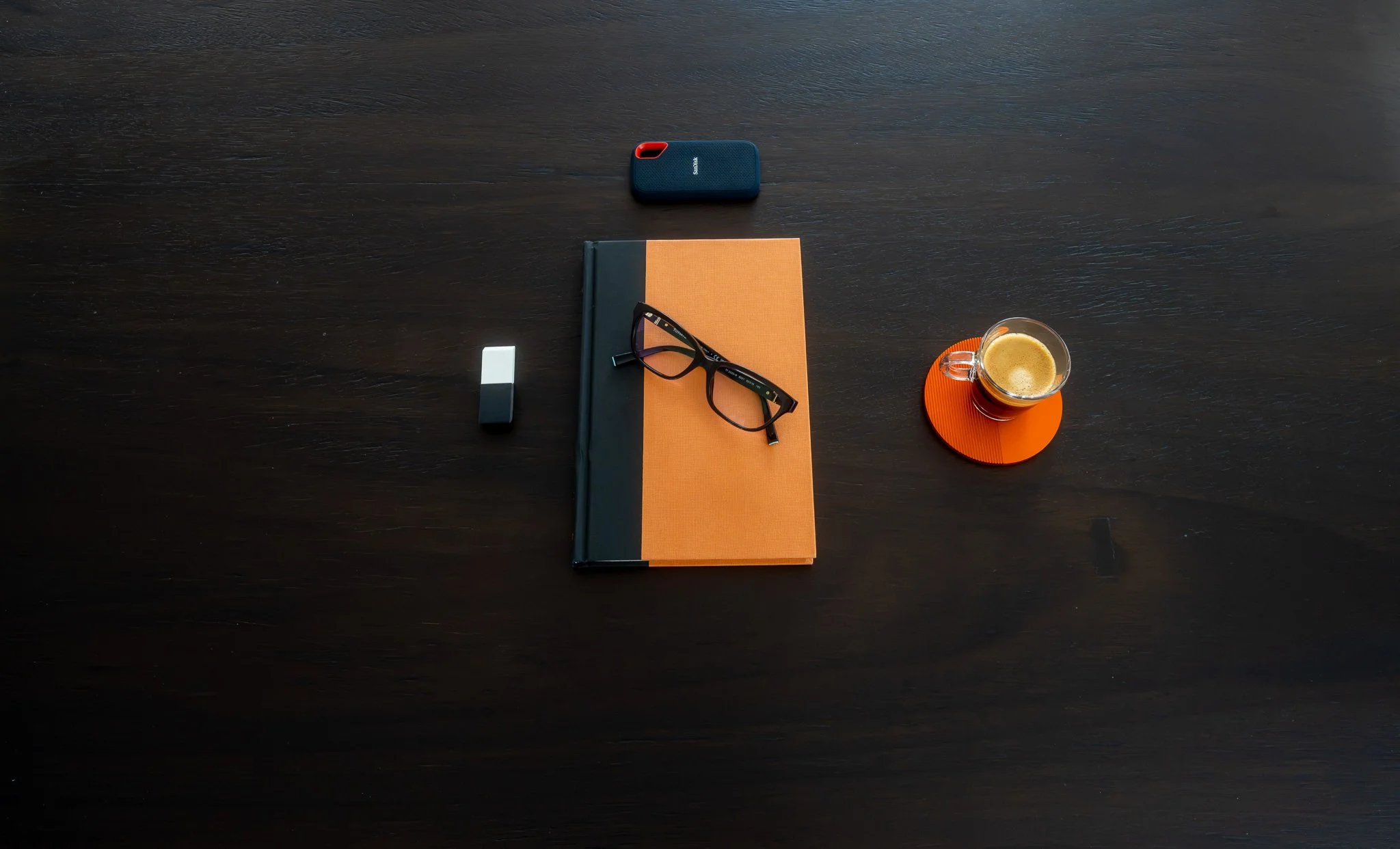A dark wooden table with a black and orange hardcover notebook, a pair of black glasses, a white eraser, a small blue and red portable speaker, and a glass mug of coffee on an orange coaster.