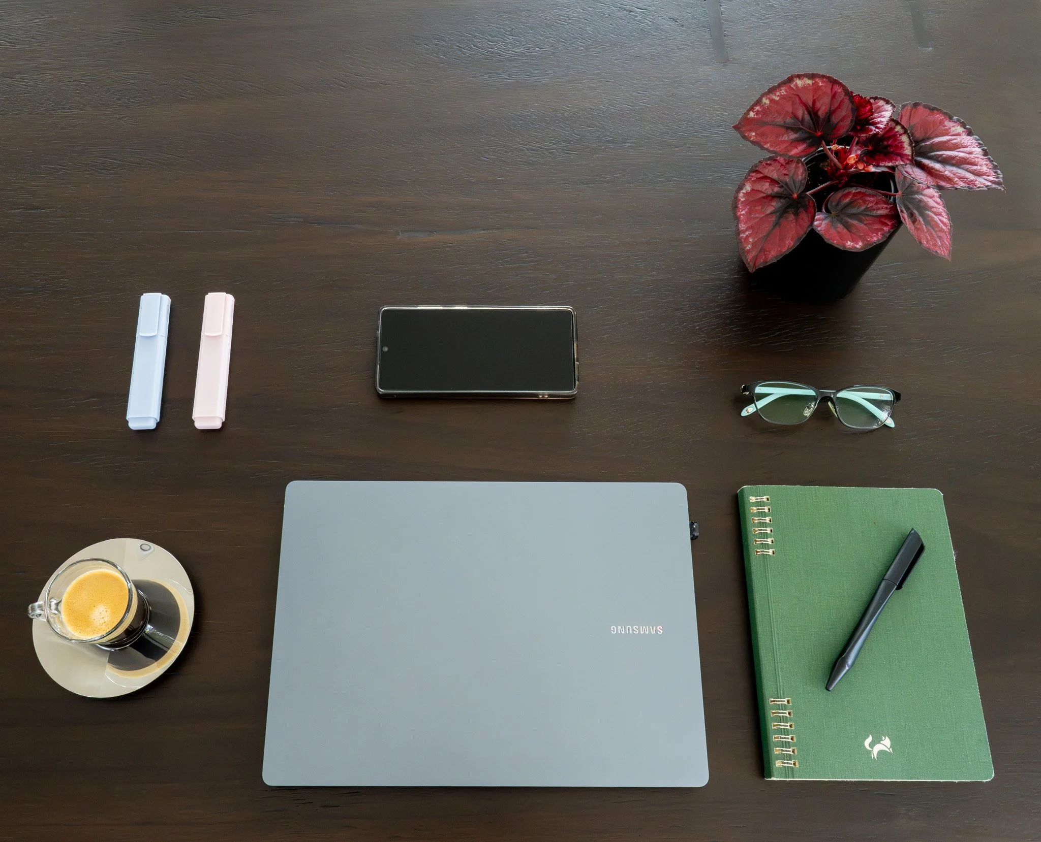 Top-down view of a wooden desk with a glass of coffee, a closed laptop, two white and pink highlighters, a green notebook with a pen, a pair of glasses, a smartphone, and a potted plant with red and green leaves.