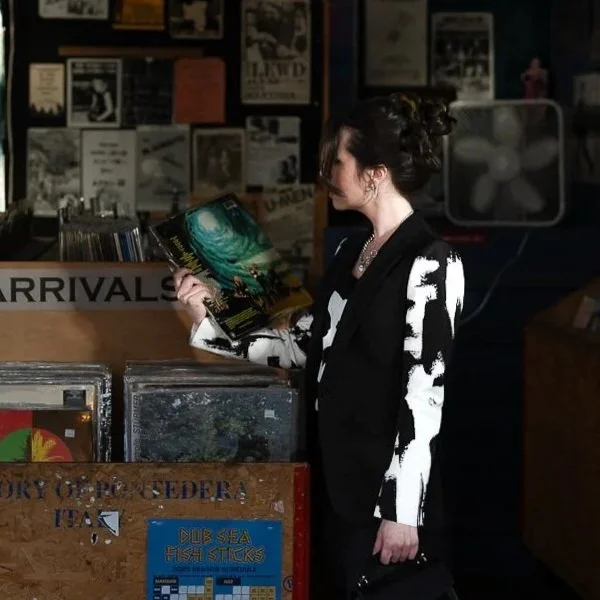 A woman with dark hair styled in an updo, wearing a black and white patterned blazer, reads a large book or magazine while standing in front of a record store or music shop. The background consists of wall displays with various posters and notices, and a fan is visible to the right.