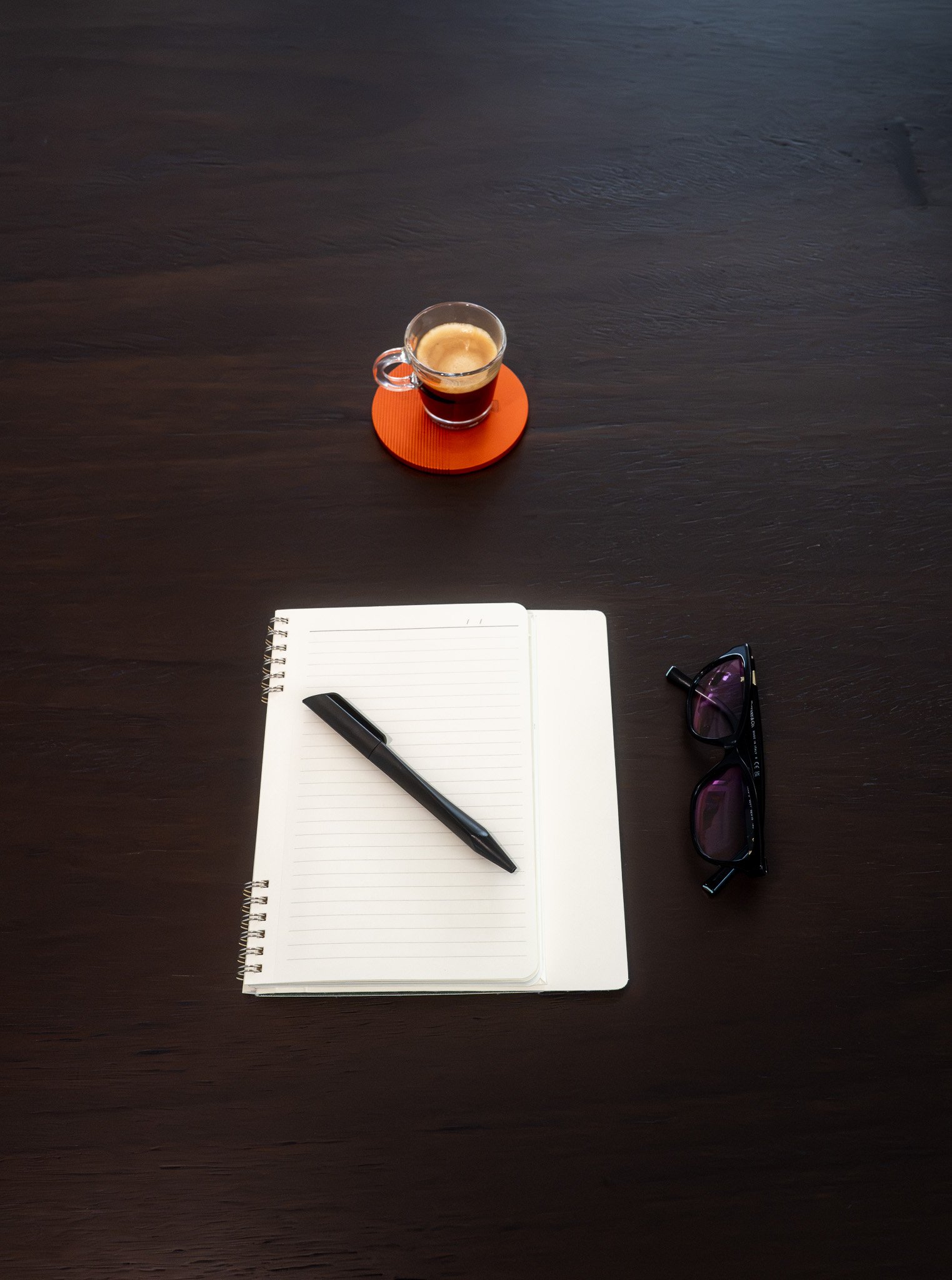 Top view of a dark wooden desk with a notebook, black pen, sunglasses, a cup of coffee on an orange coaster, and a partially visible white paper.