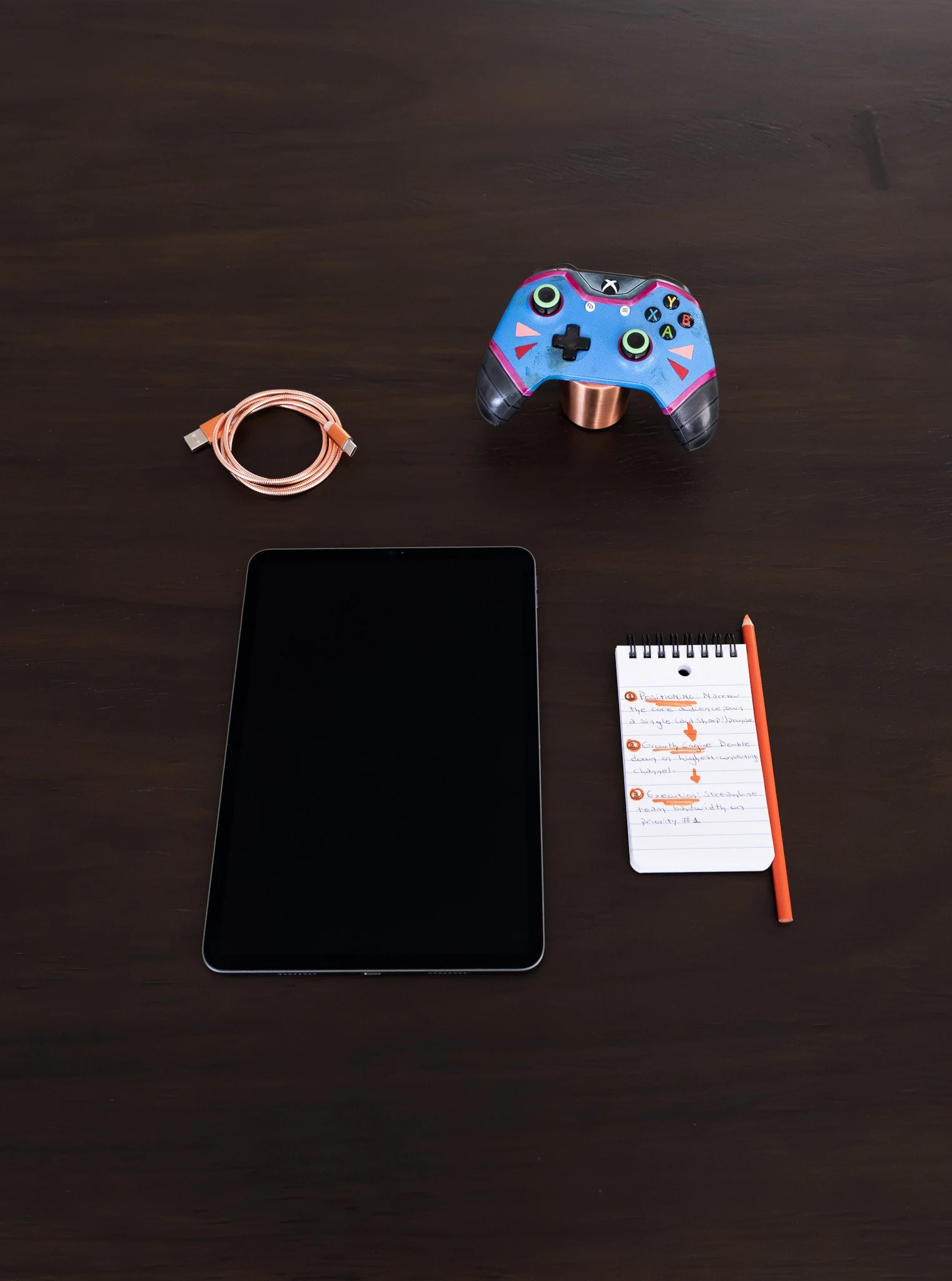 A dark wooden desk with a tablet, a colorful gaming controller, a coiled charging cable, a small notepad with handwritten notes, and an orange pen.
