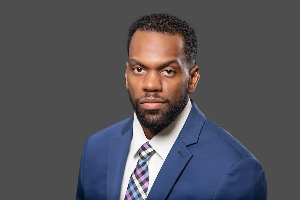Professional man with dark hair and beard wearing a blue suit, white shirt, and patterned tie, looking into the camera against a gray background.