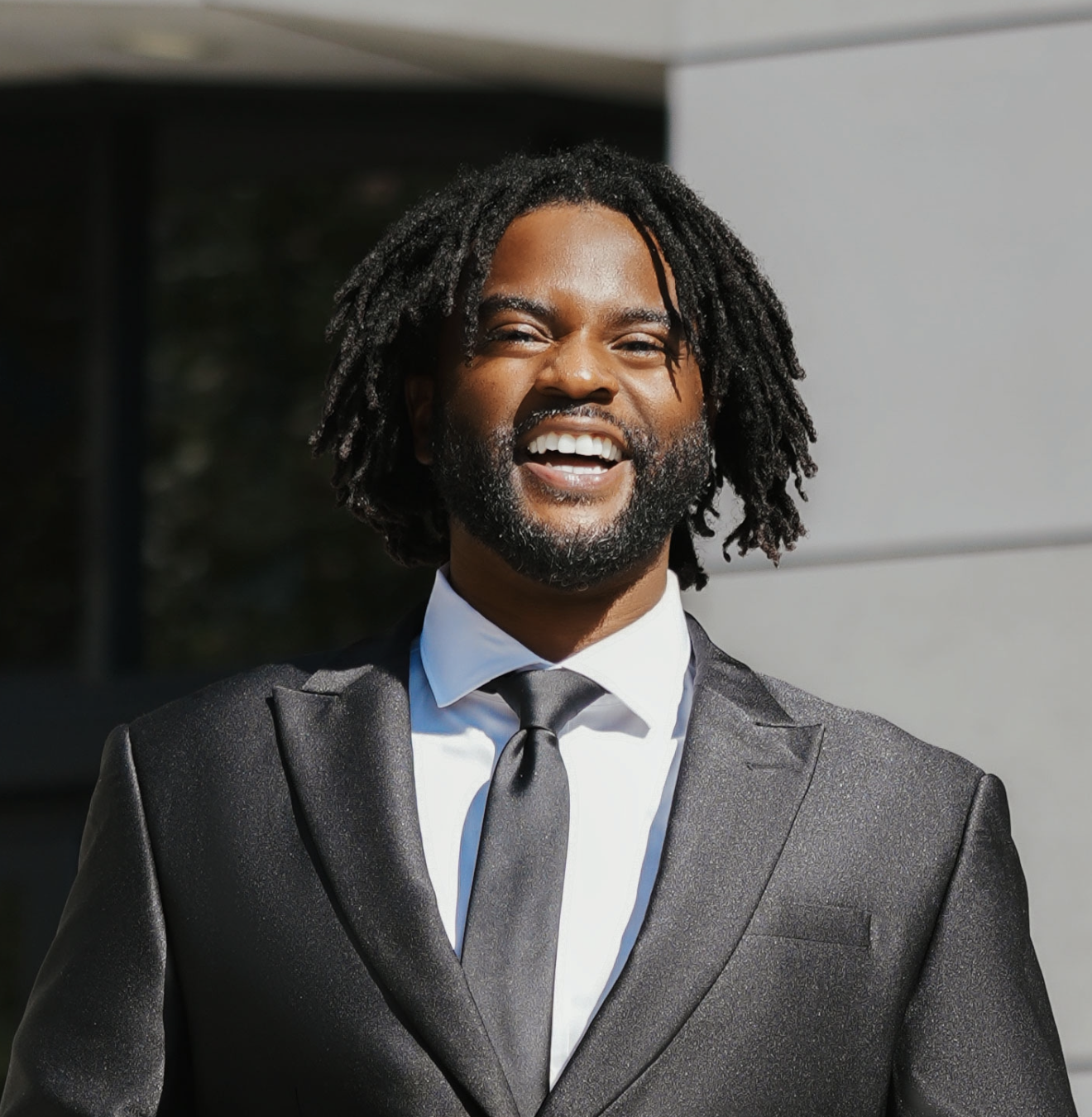 A man with dreadlocks and a beard smiling, wearing a black suit, white dress shirt, and black tie, standing outdoors in front of a modern building.