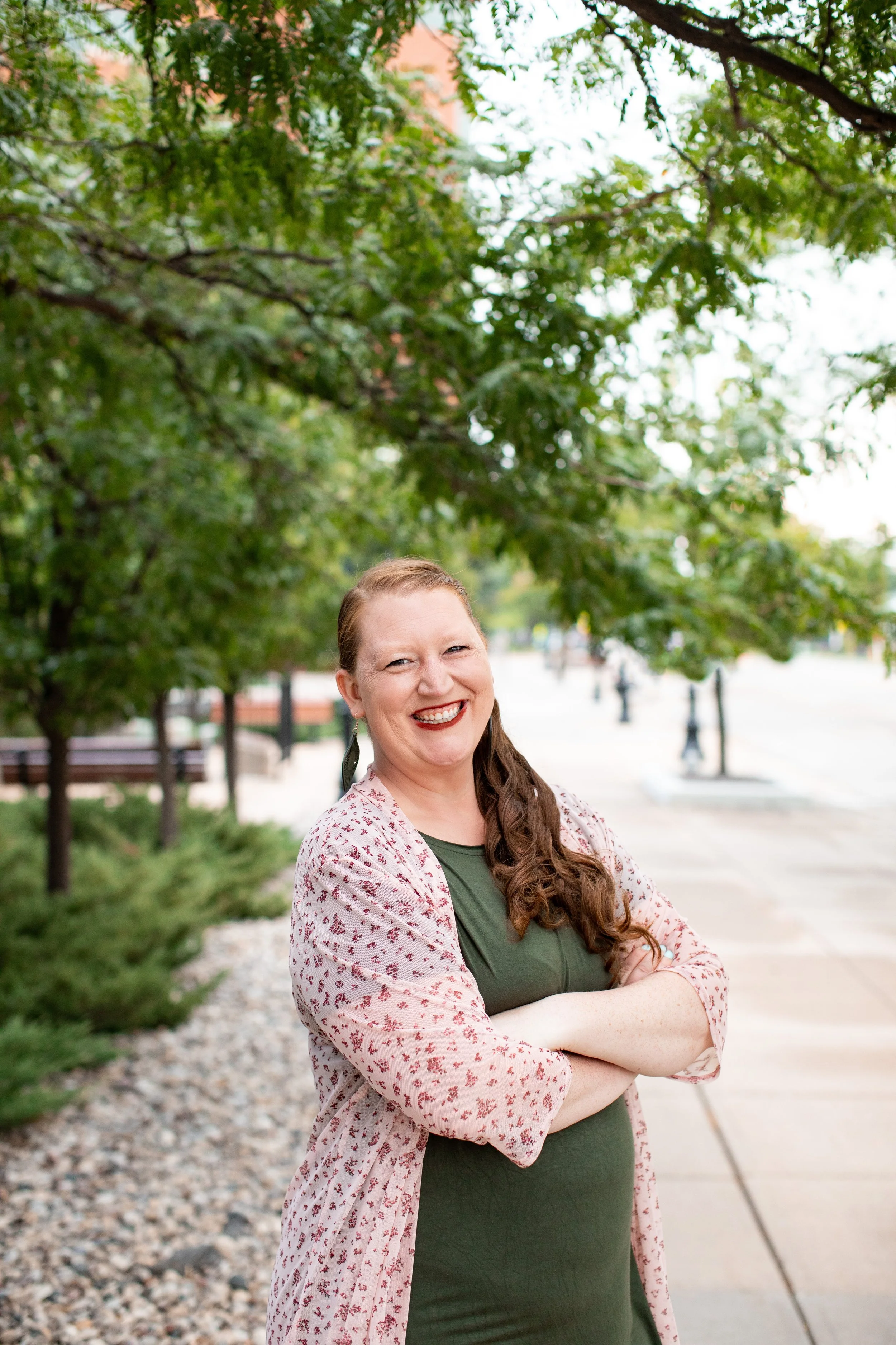 A woman with long brown hair in a braid, standing outdoors with trees and a sidewalk, smiling with arms crossed, wearing a green dress and a light pink cardigan with red patterns.
