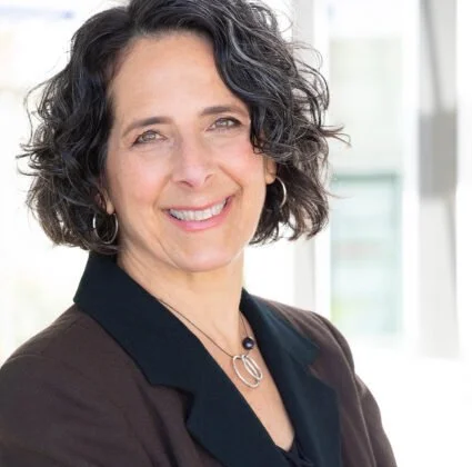 A smiling middle-aged woman with short curly dark hair, wearing a black blazer and a necklace, in a bright indoor setting with windows in the background.