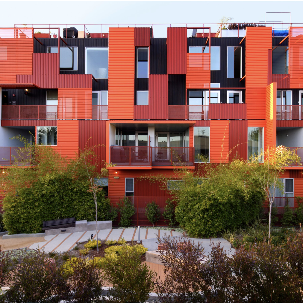 Modern multi-story apartment building with red and black exterior panels, glass balconies, and surrounding greenery.