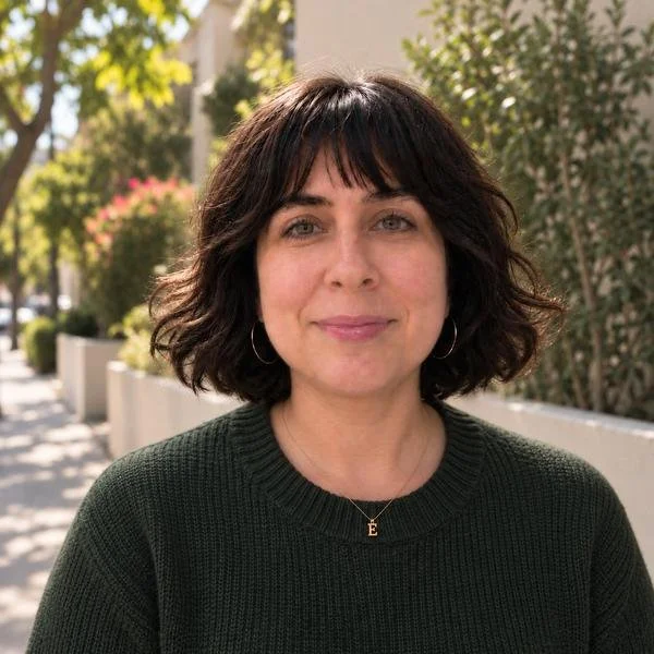 A woman with short, wavy dark hair standing outdoors in a sunny environment, wearing a dark green sweater and a gold necklace with the letter E, smiling at the camera.