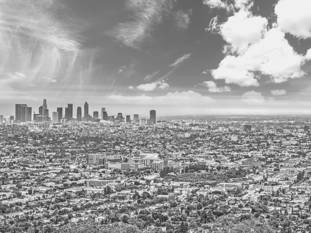 A black and white aerial view of Los Angeles, showing the downtown skyline with high-rise buildings, surrounded by a sprawling urban area, under a partly cloudy sky.