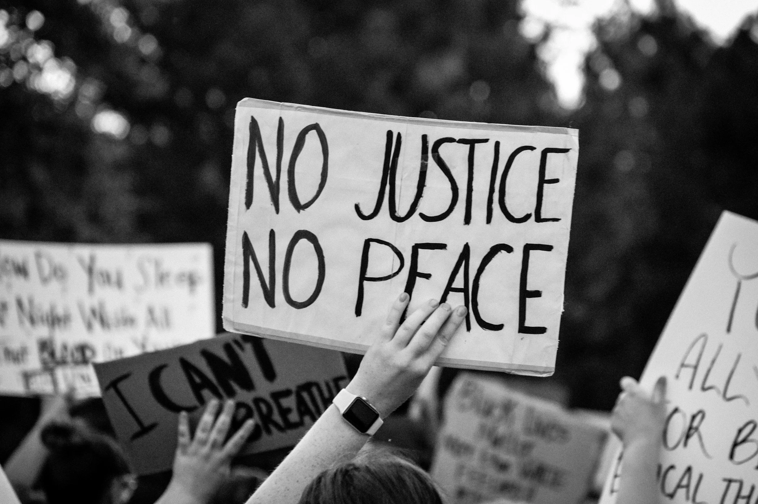 A protest scene with a person holding a sign that says "NO JUSTICE NO PEACE" among other signs, in black and white.