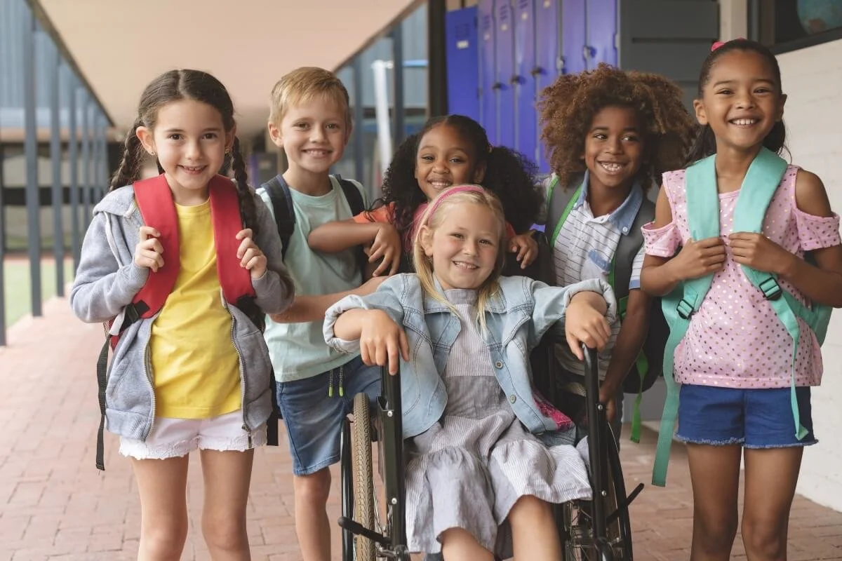 Group of six diverse children, five standing and one in a wheelchair, smiling at school hallway.