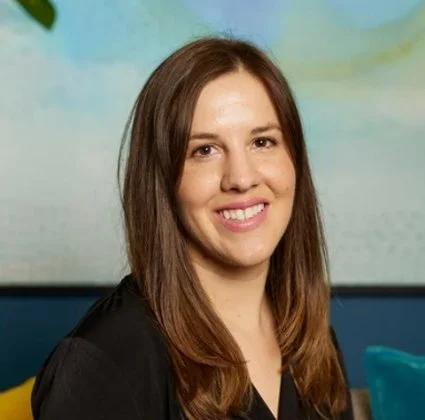 A young woman with long brown hair smiling at the camera, sitting on a colorful couch with a green and blue background.