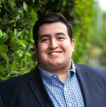 Portrait of a smiling man wearing a navy blazer and blue checked shirt outdoors between green bushes.