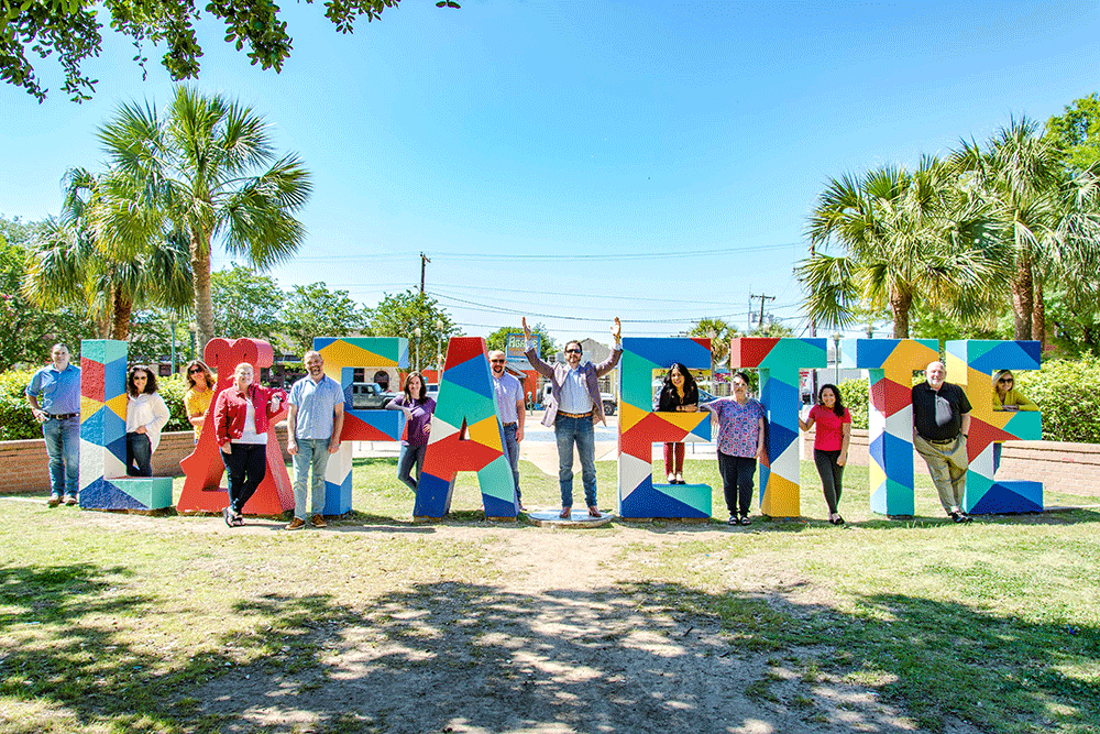 People posing in front of colorful 'LAKE' sign outdoors under palm trees on sunny day.