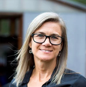 A woman with blonde hair and glasses smiling outdoors.