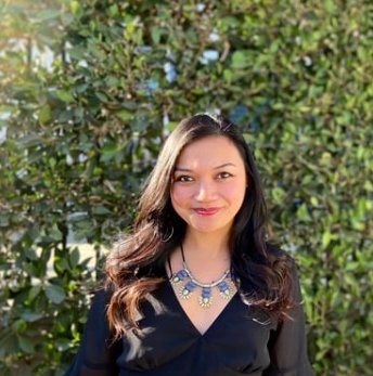 A woman with long brown hair, smiling, wearing a black top and a statement necklace, standing outdoors in front of green foliage.