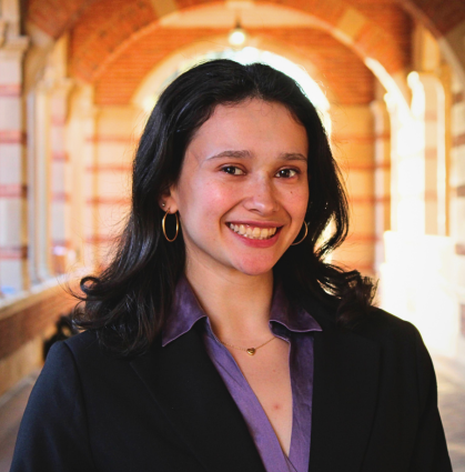 Smiling woman with dark hair, wearing a black blazer and purple shirt, in a bright, arched brick hallway.